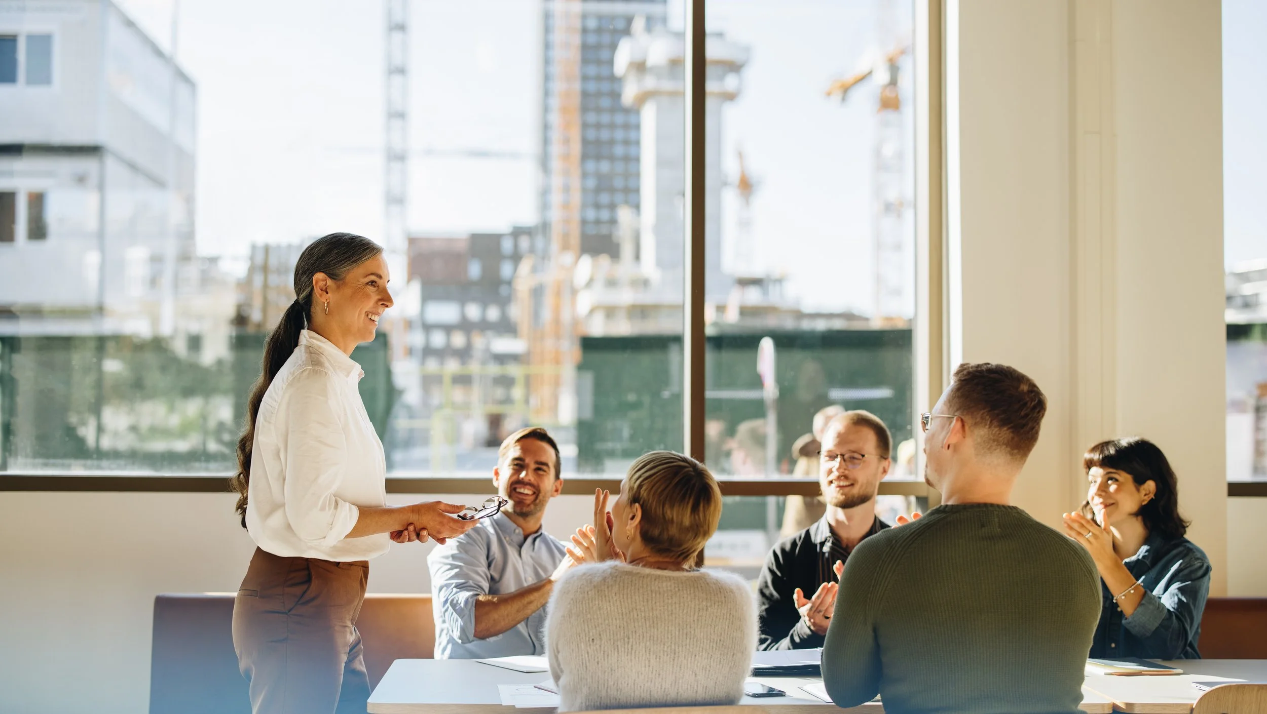 A diverse group of six people, five sitting at a table and one standing, in a meeting room with large windows showing a cityscape outside. The woman standing appears to be speaking or presenting, while the others are smiling and clapping.