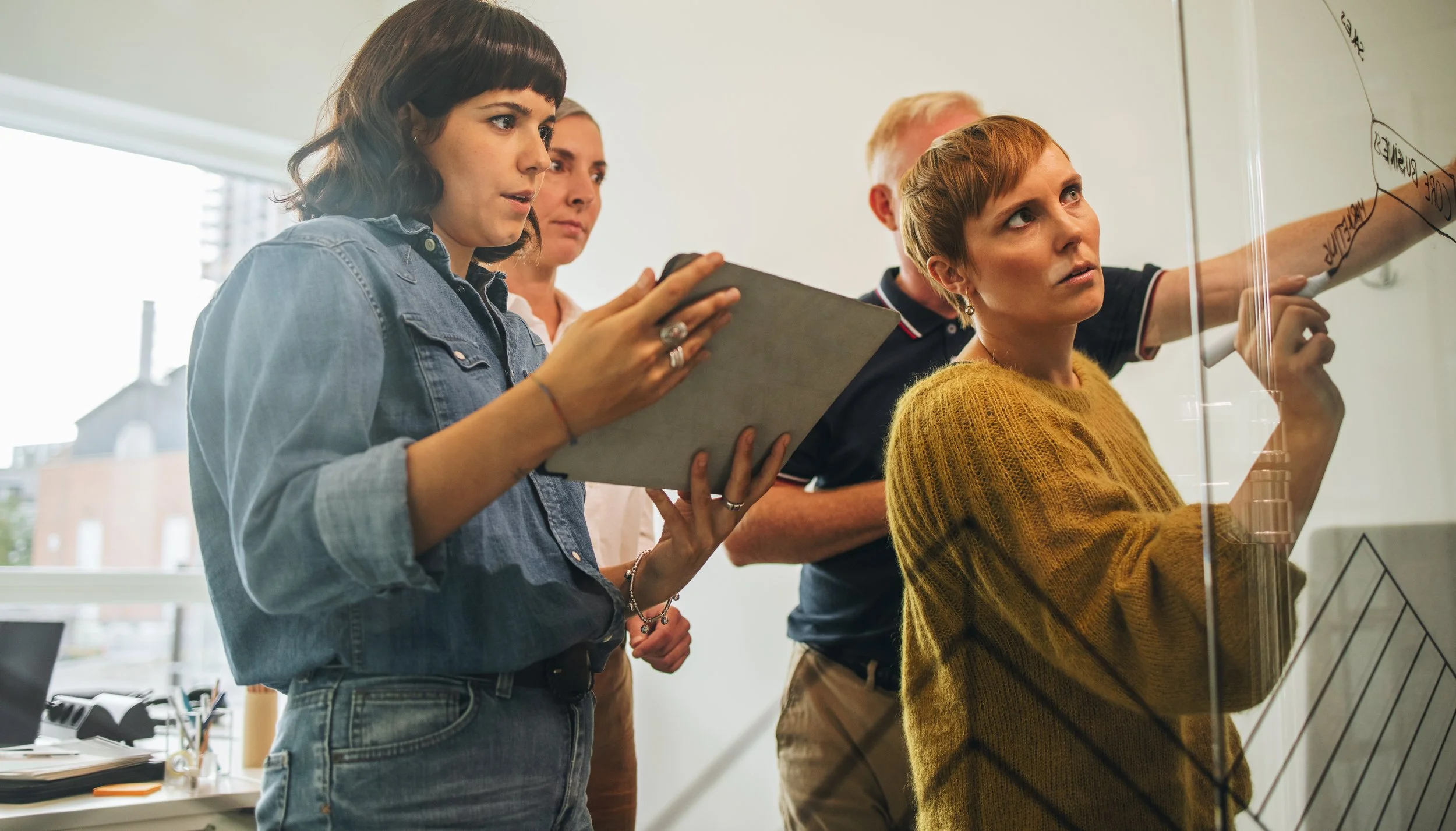 Four people in casual clothing collaborating and analyzing ideas on a whiteboard in a modern office.