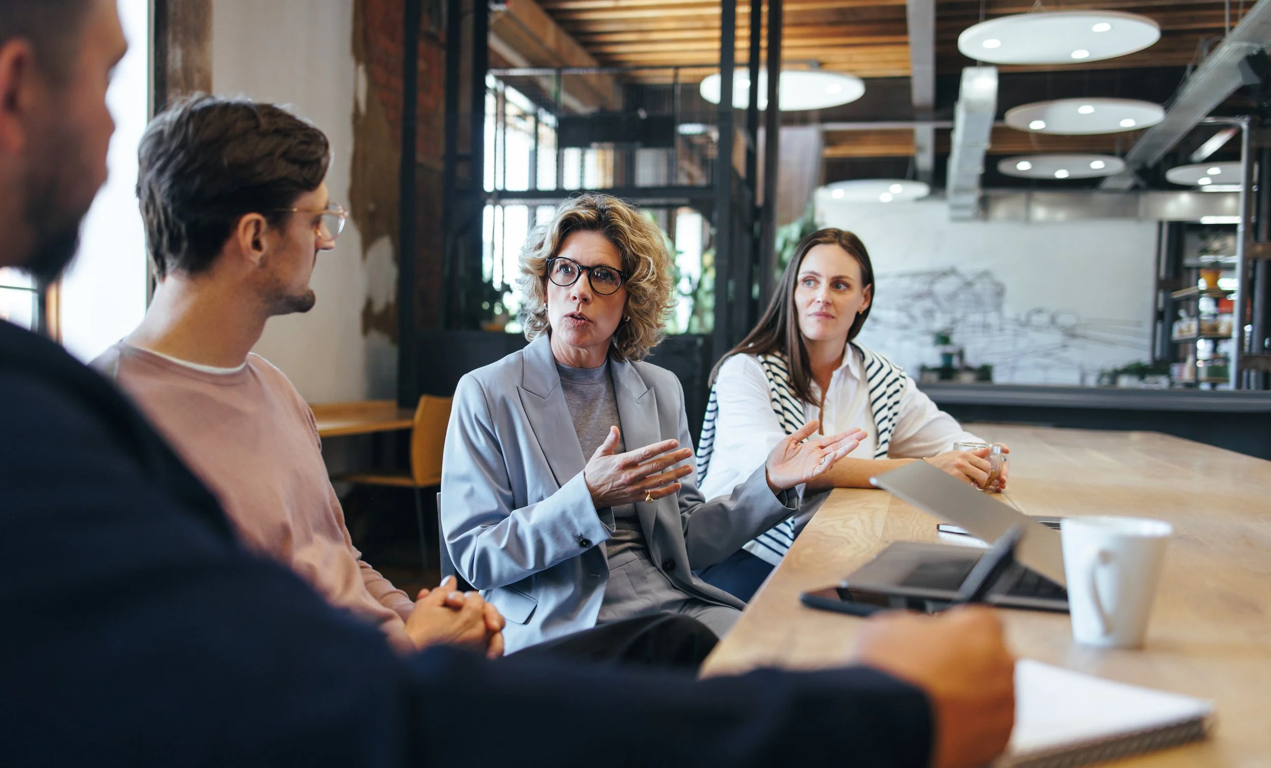 Group of four people having a discussion at a wooden table in a modern cafe or office space.