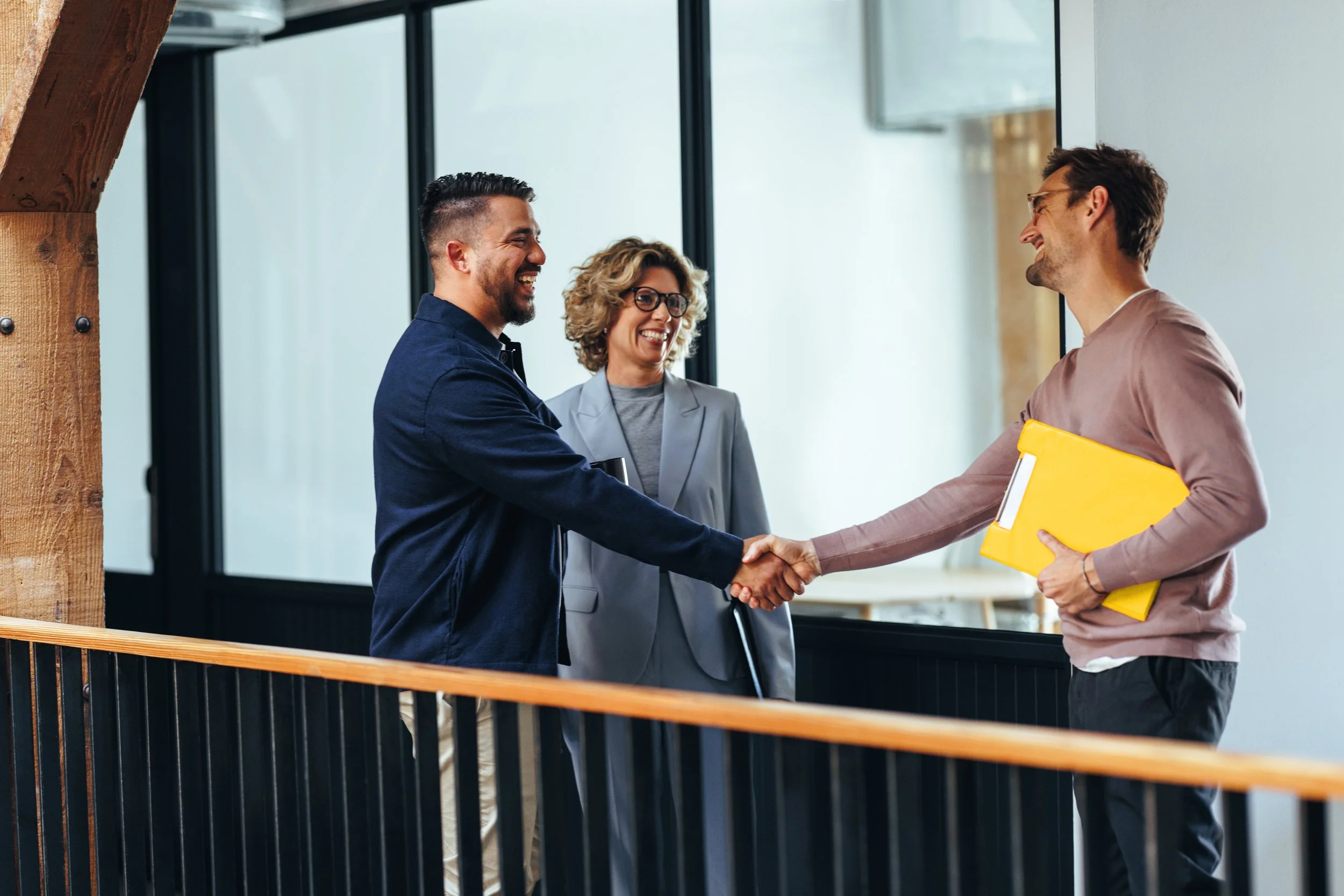 Two men shaking hands in an office setting while a woman watches and smiles.