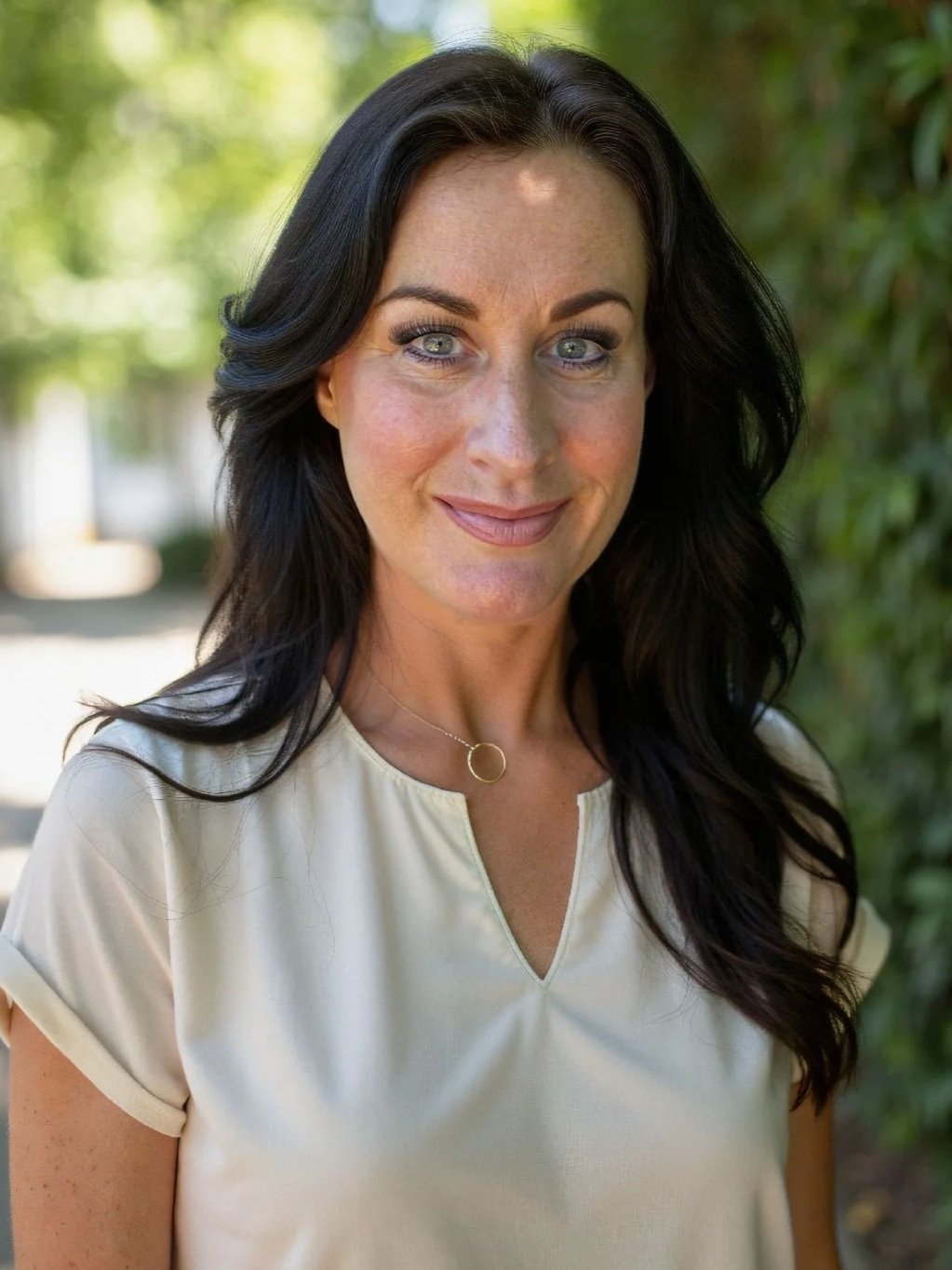 A woman with long dark hair, blue eyes, and light skin wearing a cream-colored top and a silver necklace, standing outdoors near greenery.