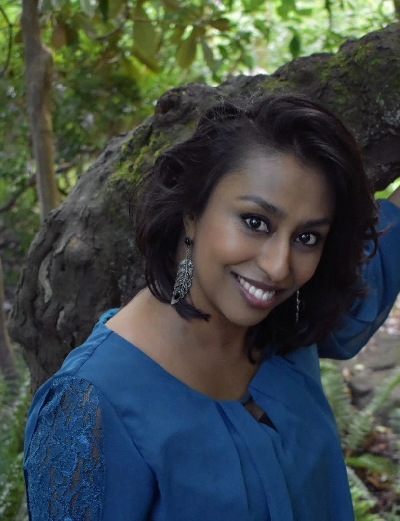 Smiling woman with dark hair, wearing a blue top and leaf-shaped earrings, leaning against a tree in a green outdoor setting.
