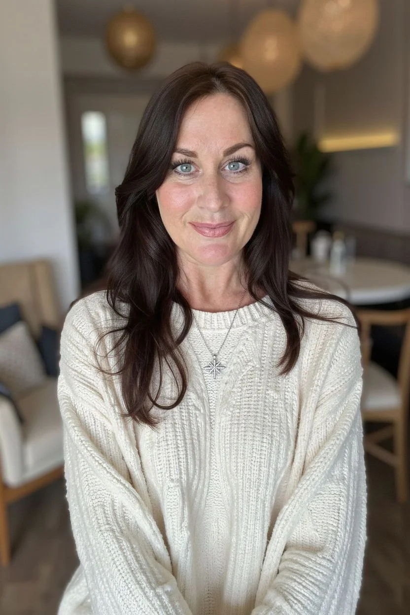 Woman with shoulder-length dark brown hair, blue eyes, and light skin, wearing a cream-colored knitted sweater and a star-shaped pendant necklace, standing indoors with blurred background of modern decor.