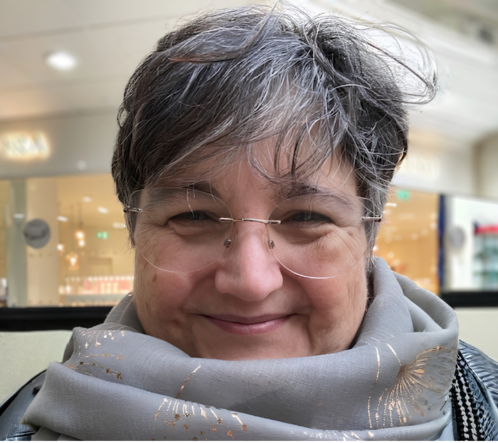 A smiling middle-aged person with short gray hair, glasses, and a light gray scarf with golden patterns, sitting in a bright indoor setting.