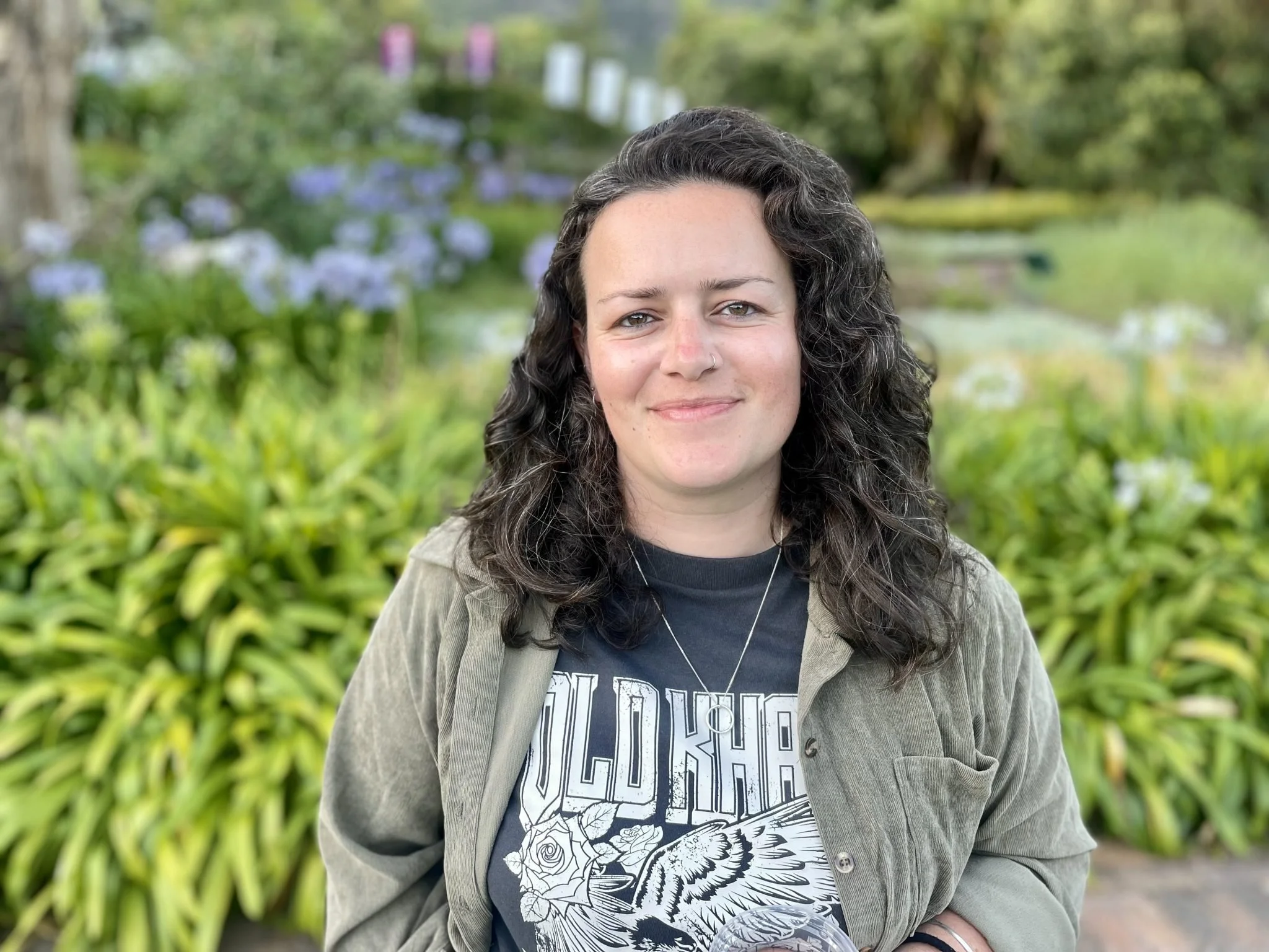 A woman with curly dark hair, light skin, and a nose piercing, smiling outdoors in front of greenery and purple flowers, wearing a graphic t-shirt and a light-colored jacket.