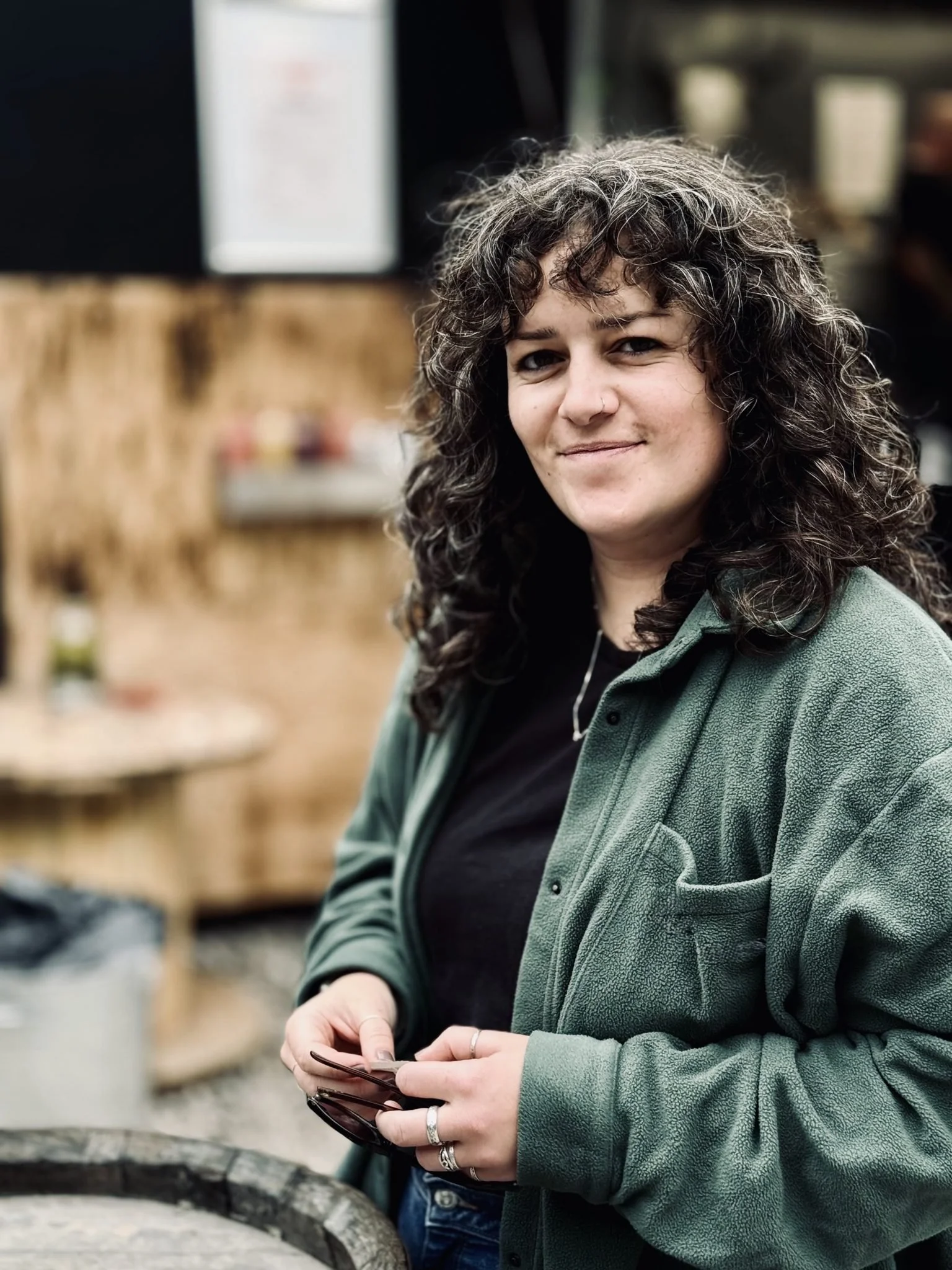 A woman with curly gray hair, wearing a green jacket and holding glasses, standing outdoors near a wooden counter, with blurred background of store shelves.