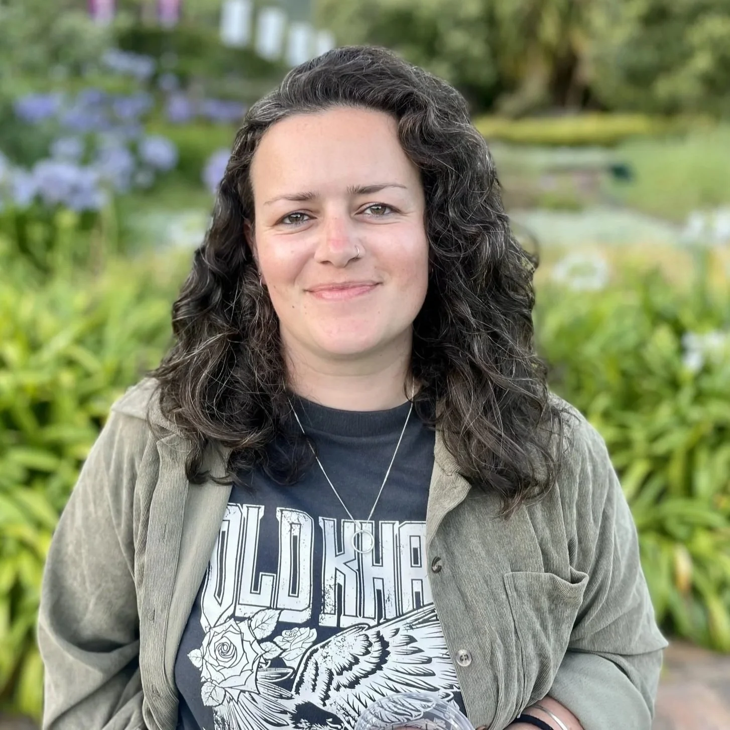A young woman with curly dark hair, light skin, and a nose piercing, smiling outdoors with green plants and purple flowers in the background, wearing a black graphic t-shirt and a gray jacket.