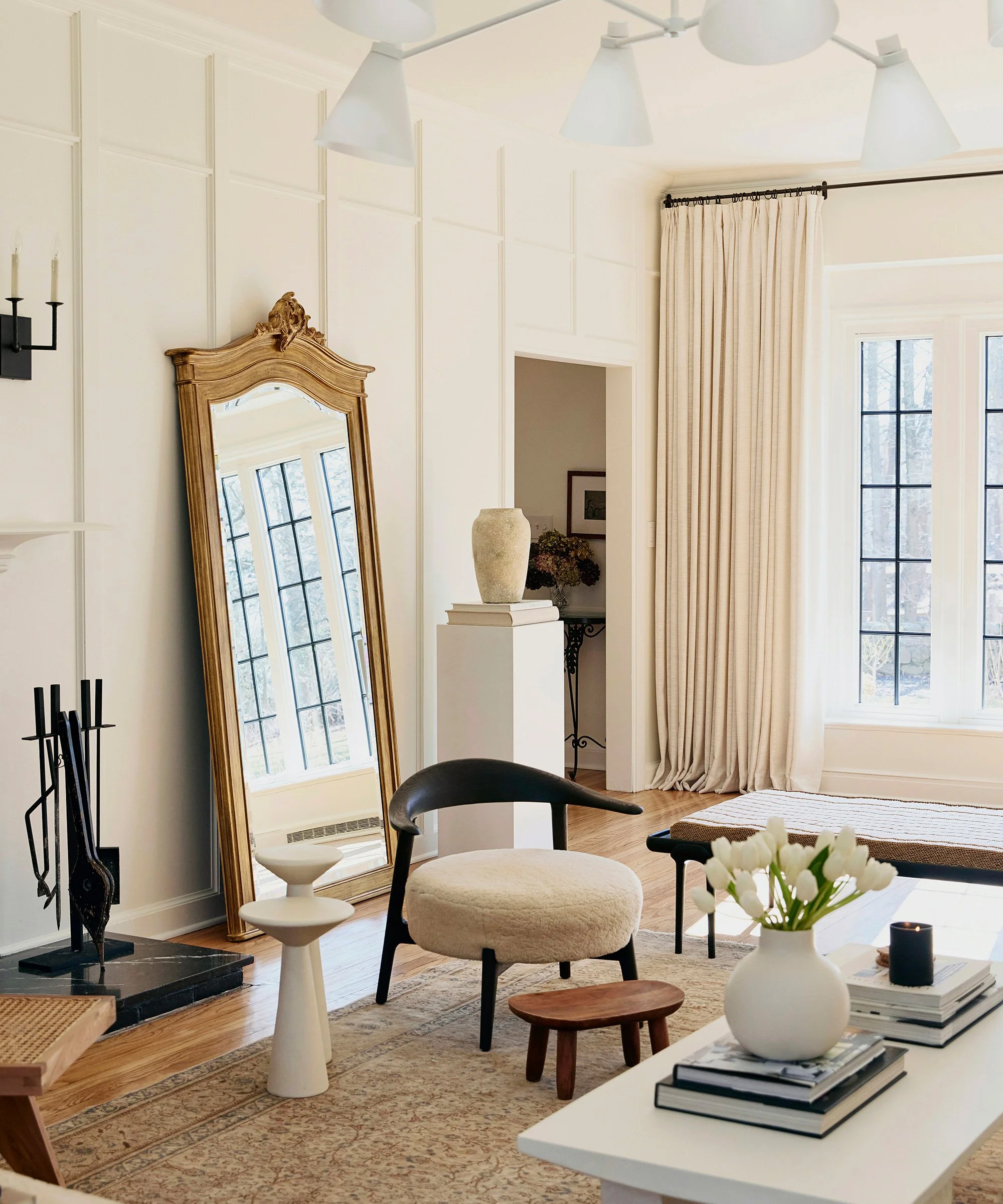 Living room with a large window, cream curtains, a full-length mirror leaning against the wall, a white chair with a cream cushion, a small wooden stool, a white table with a vase of white tulips, and a fireplace with a wood and metal holder.