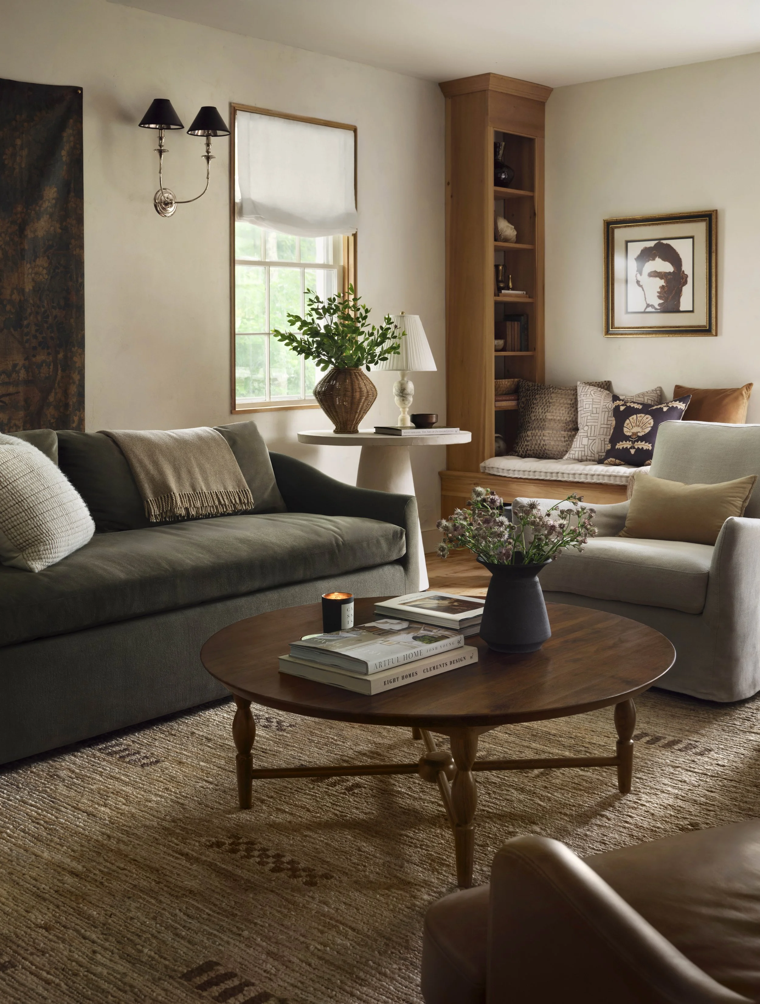 Cozy living room with a gray sofa, white armchair, wooden coffee table with books and a flower vase, window with a white curtain, wall artwork, bookshelf, and decorative pillows.