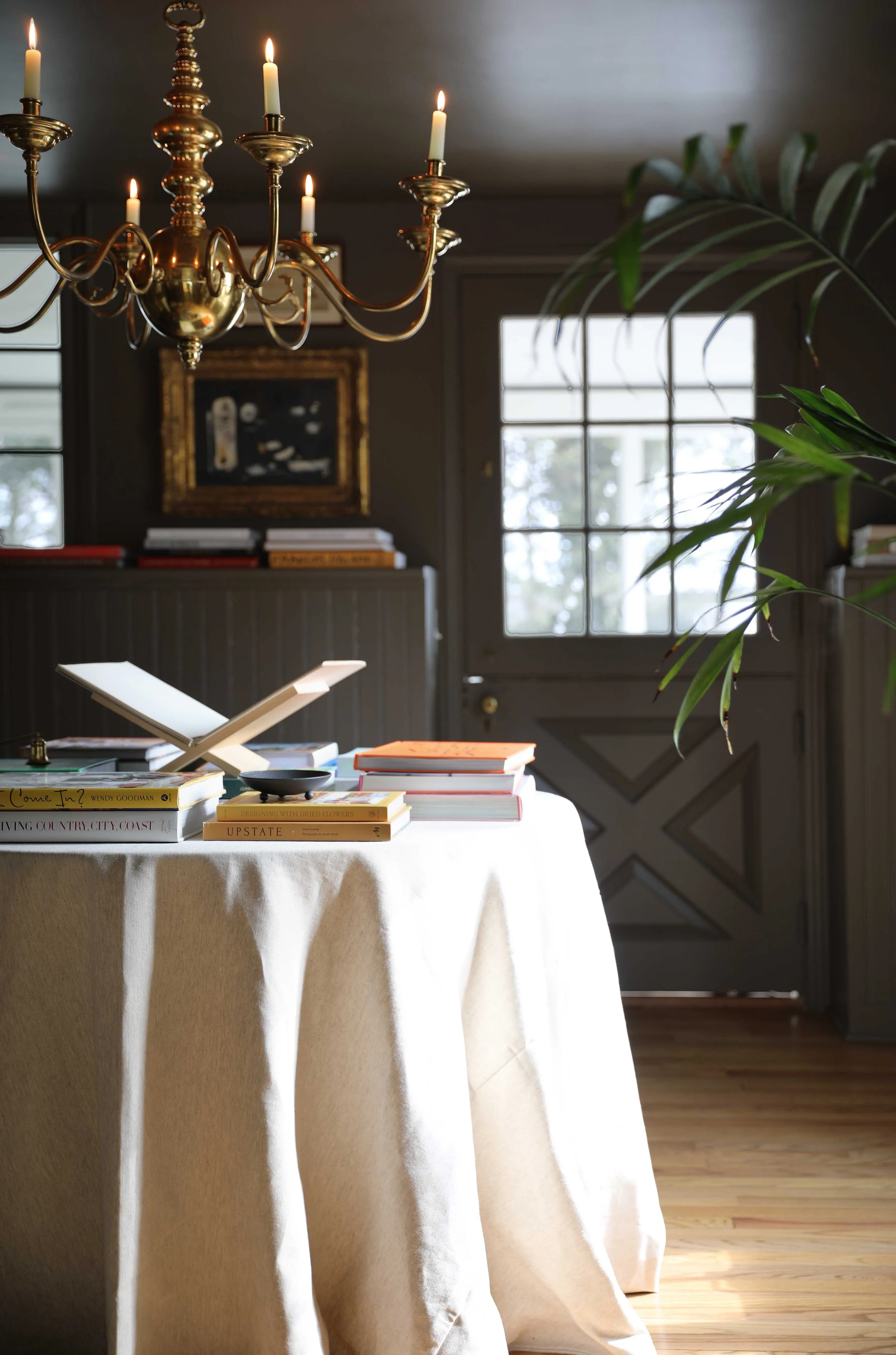 A round table covered with a white tablecloth, books, and a book stand in a warmly lit room with a chandelier, window, artwork, and greenery.