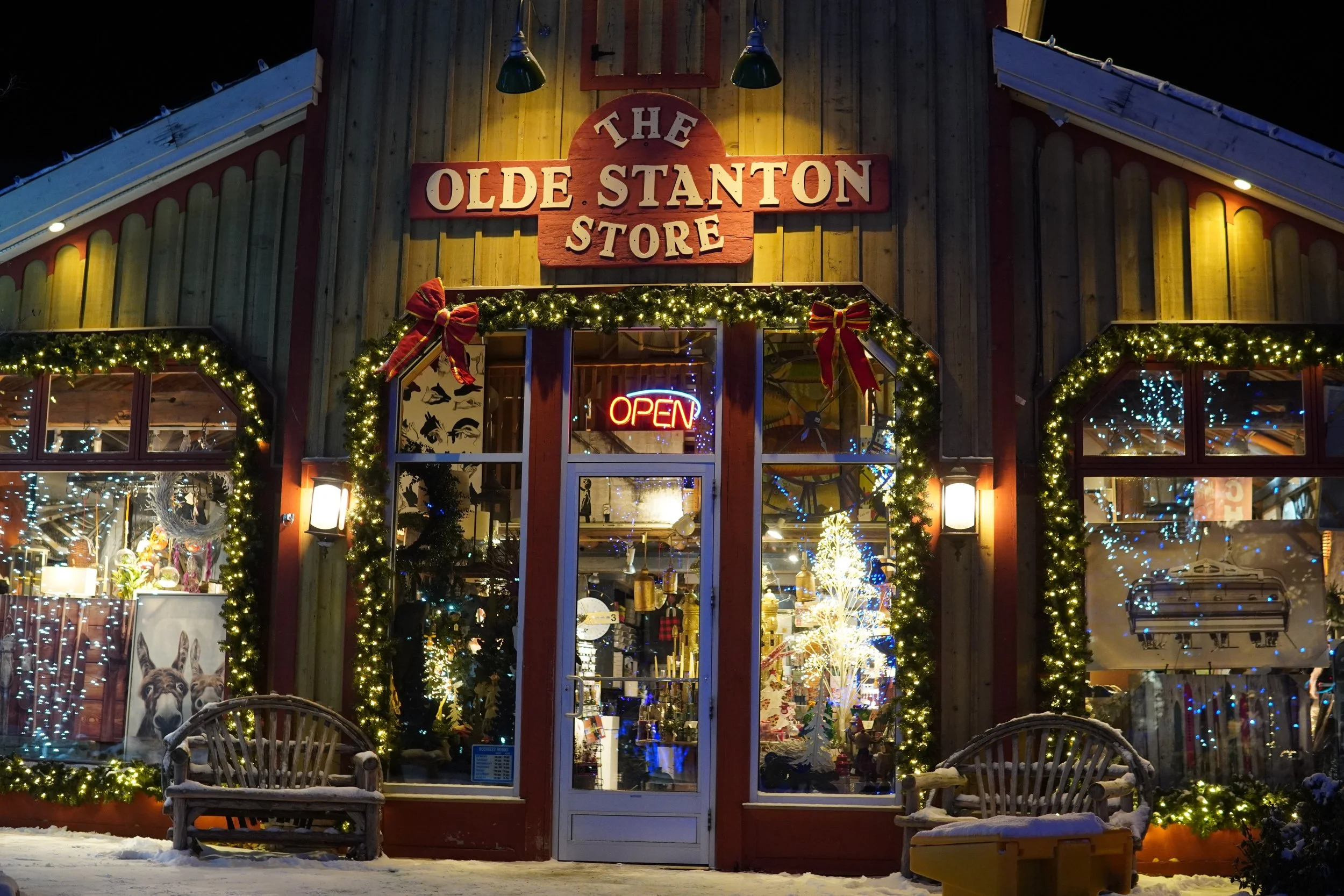 A store decorated with Christmas lights and garlands, with a sign that says 'The Olde Stanton Store' above the entrance. The storefront has large windows, and inside there are Christmas decorations and holiday-themed items visible. There are two benc
