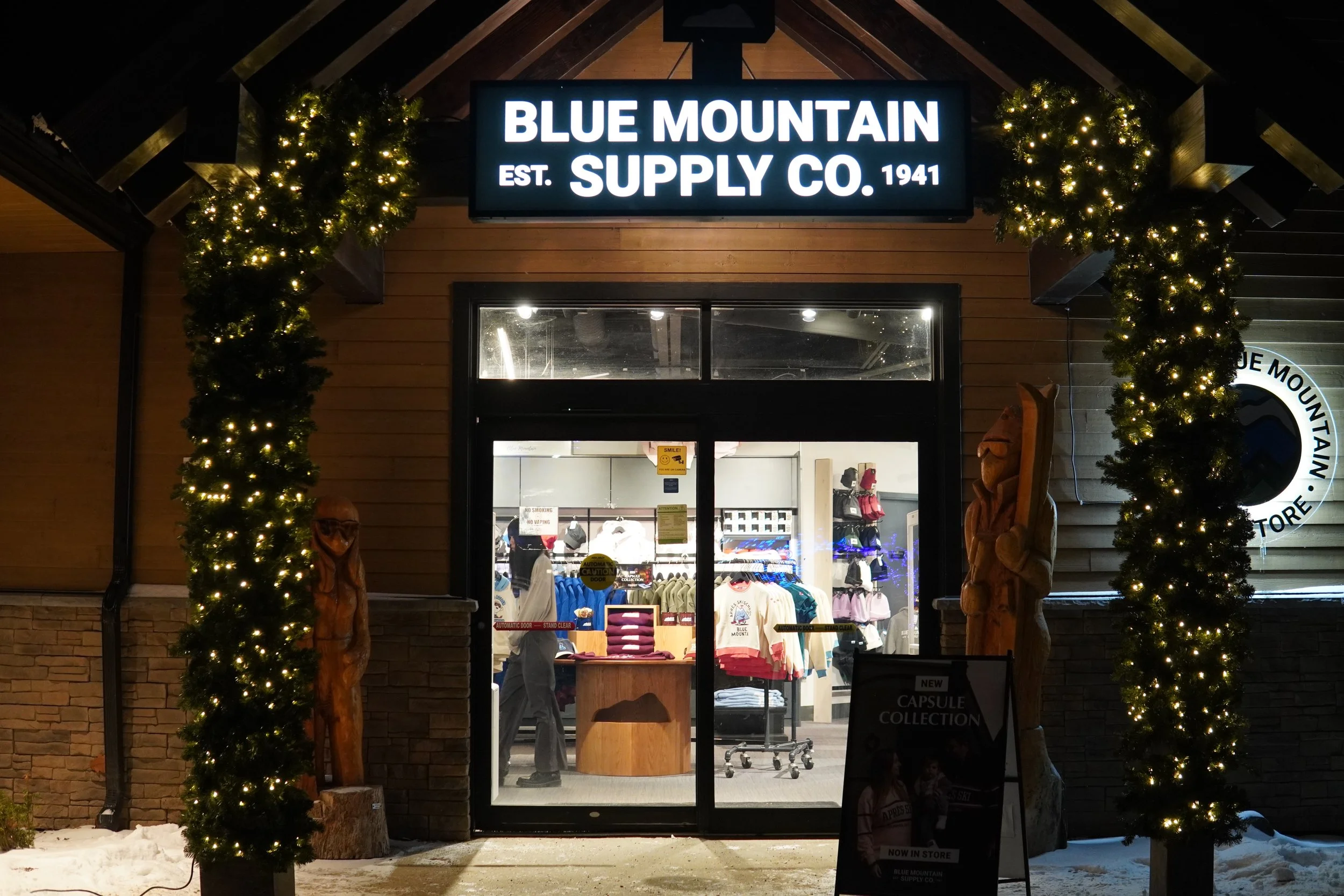Storefront of Blue Mountain Supply Co. with a illuminated sign, decorated with string lights on pillars, and featuring wooden carvings on each side of the entrance, with snow on the ground outside.