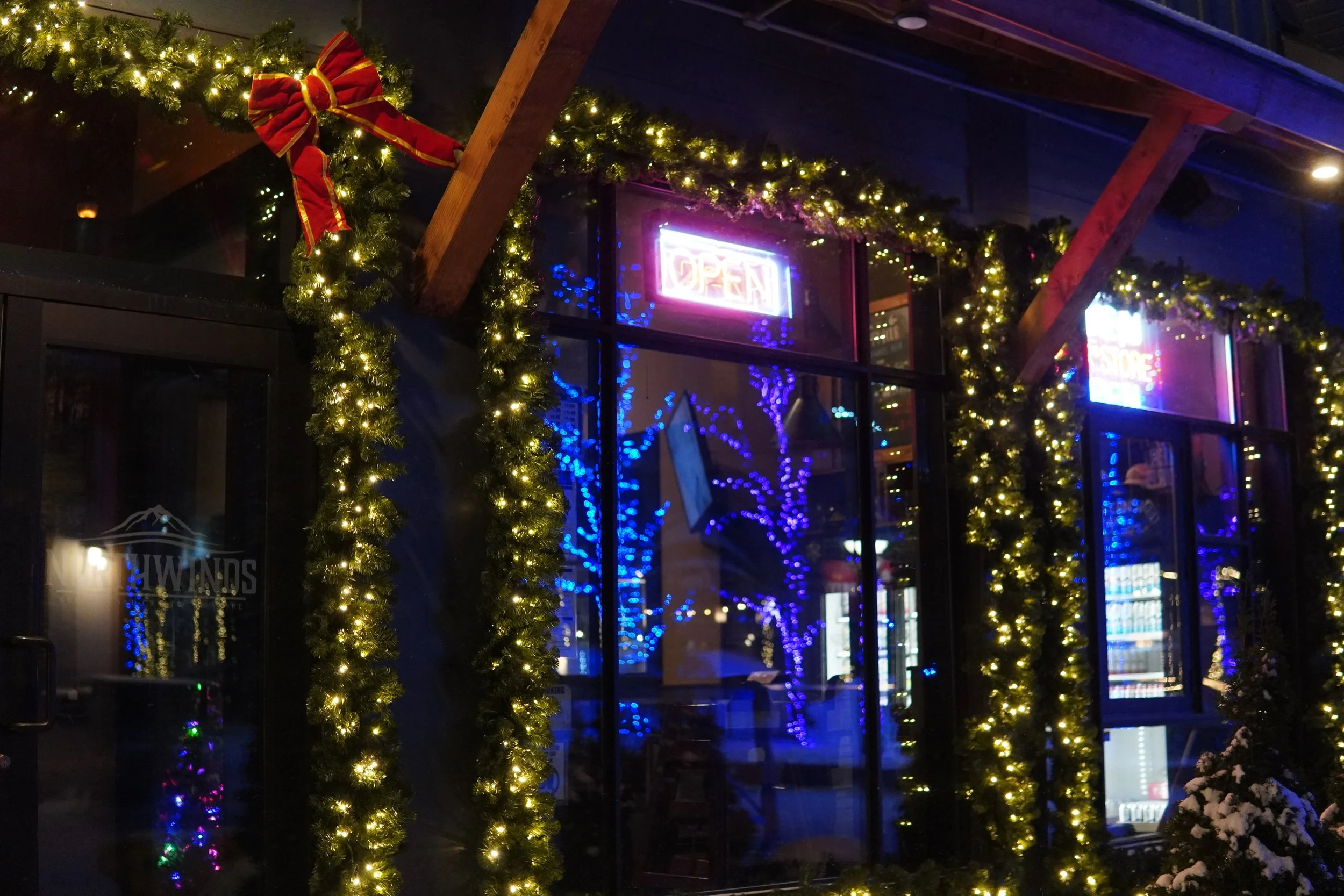 The exterior of a store decorated with Christmas lights and garlands, with a neon open sign in the window at night.