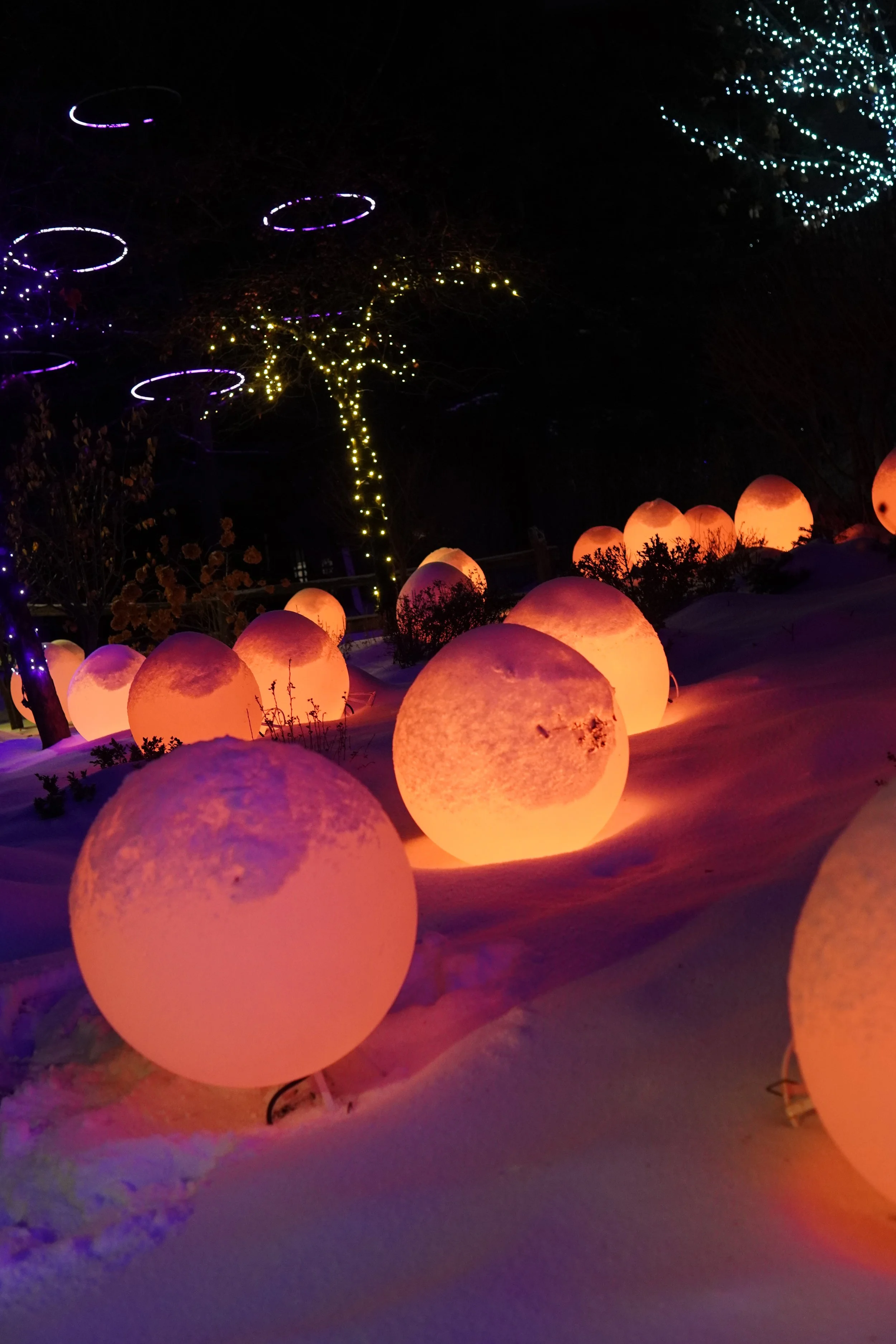 Snow-covered ground with illuminated orange spherical sculptures, decorated trees with string lights, and glowing circular light fixtures in the night sky.