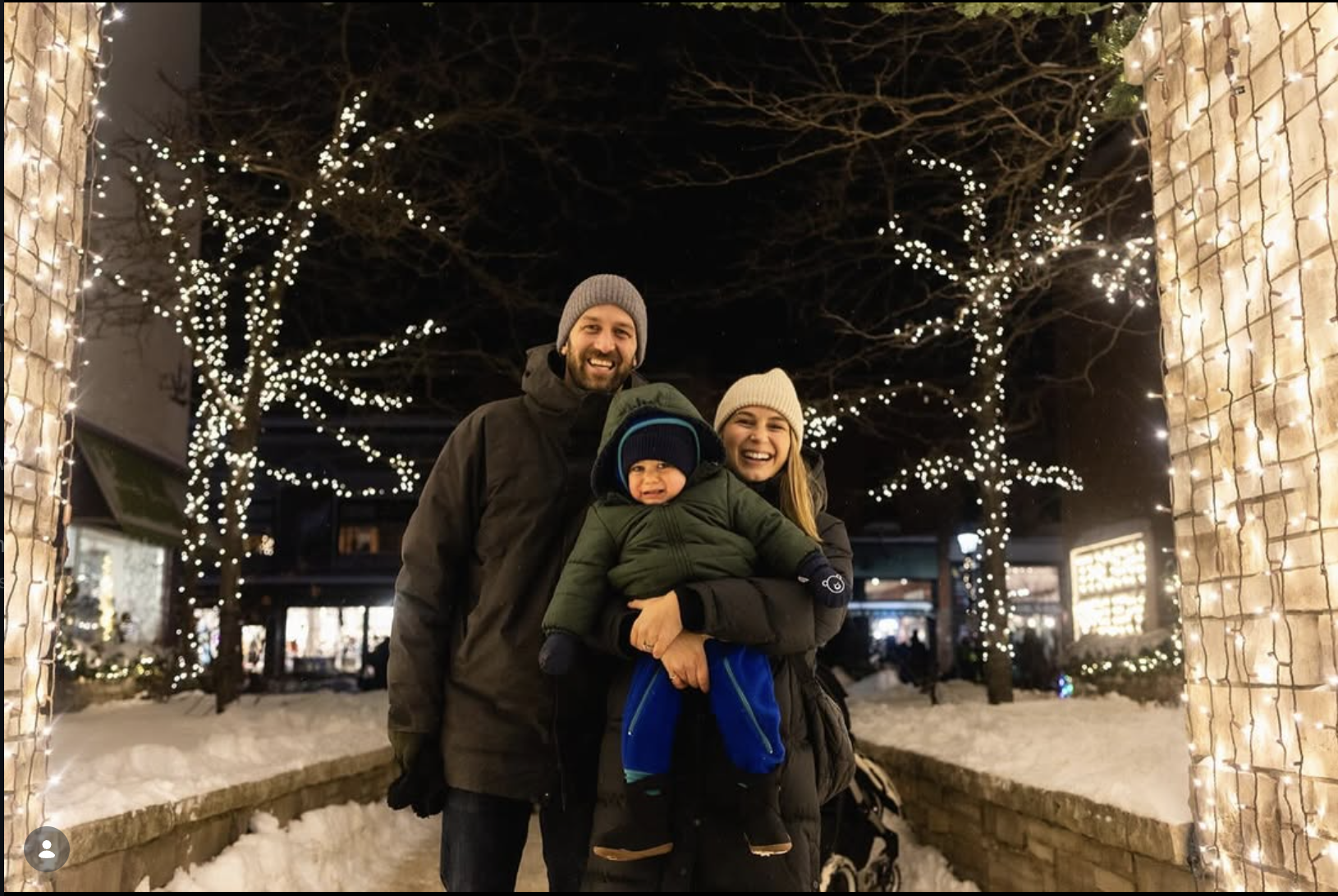 A family of three, with a man, woman, and a small child, standing outdoors at night under Christmas lights and decorations, smiling and posing for a photo in a festive winter setting with snow on the ground.