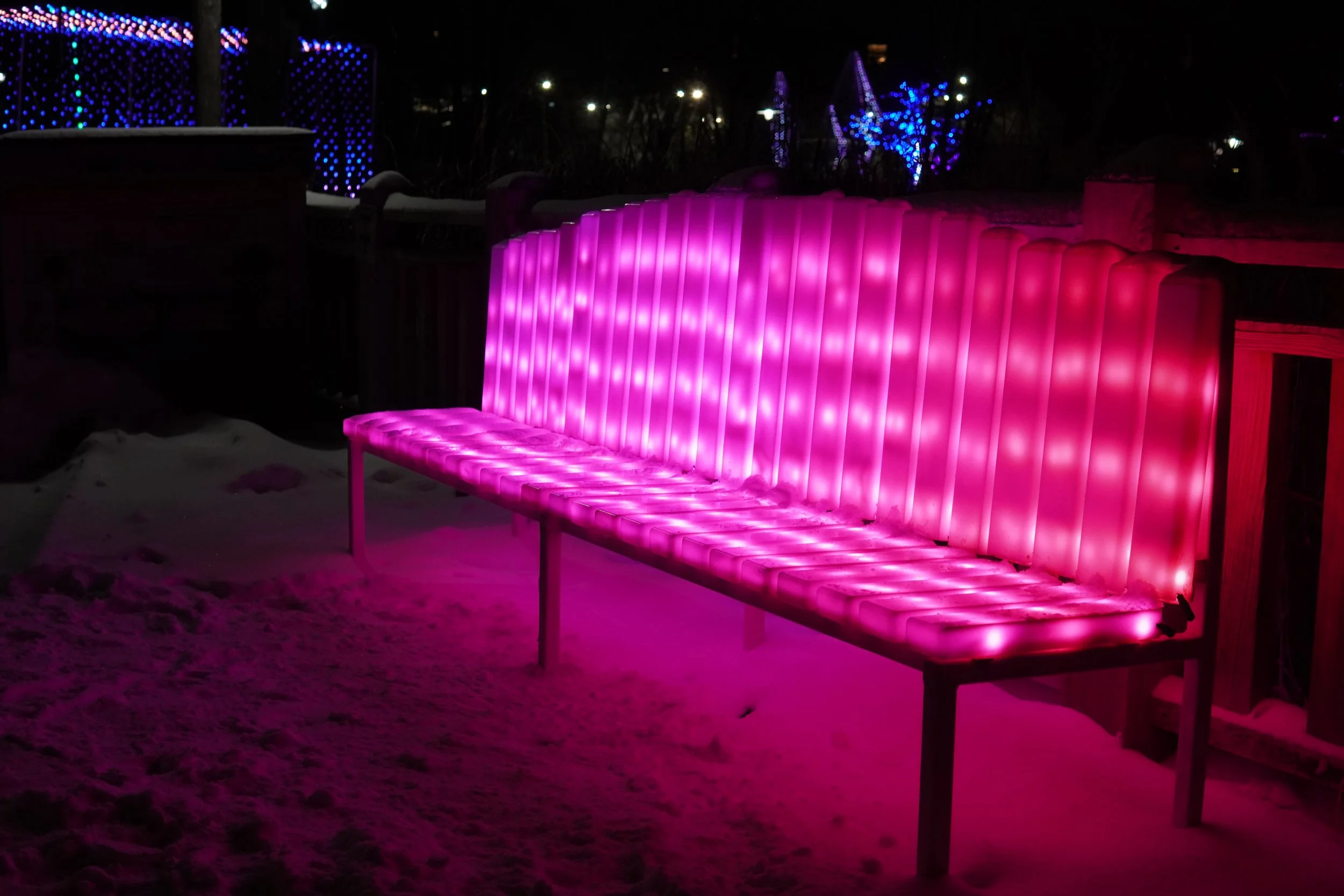 Illuminated pink outdoor bench at night with snow on the ground and colorful string lights in the background.