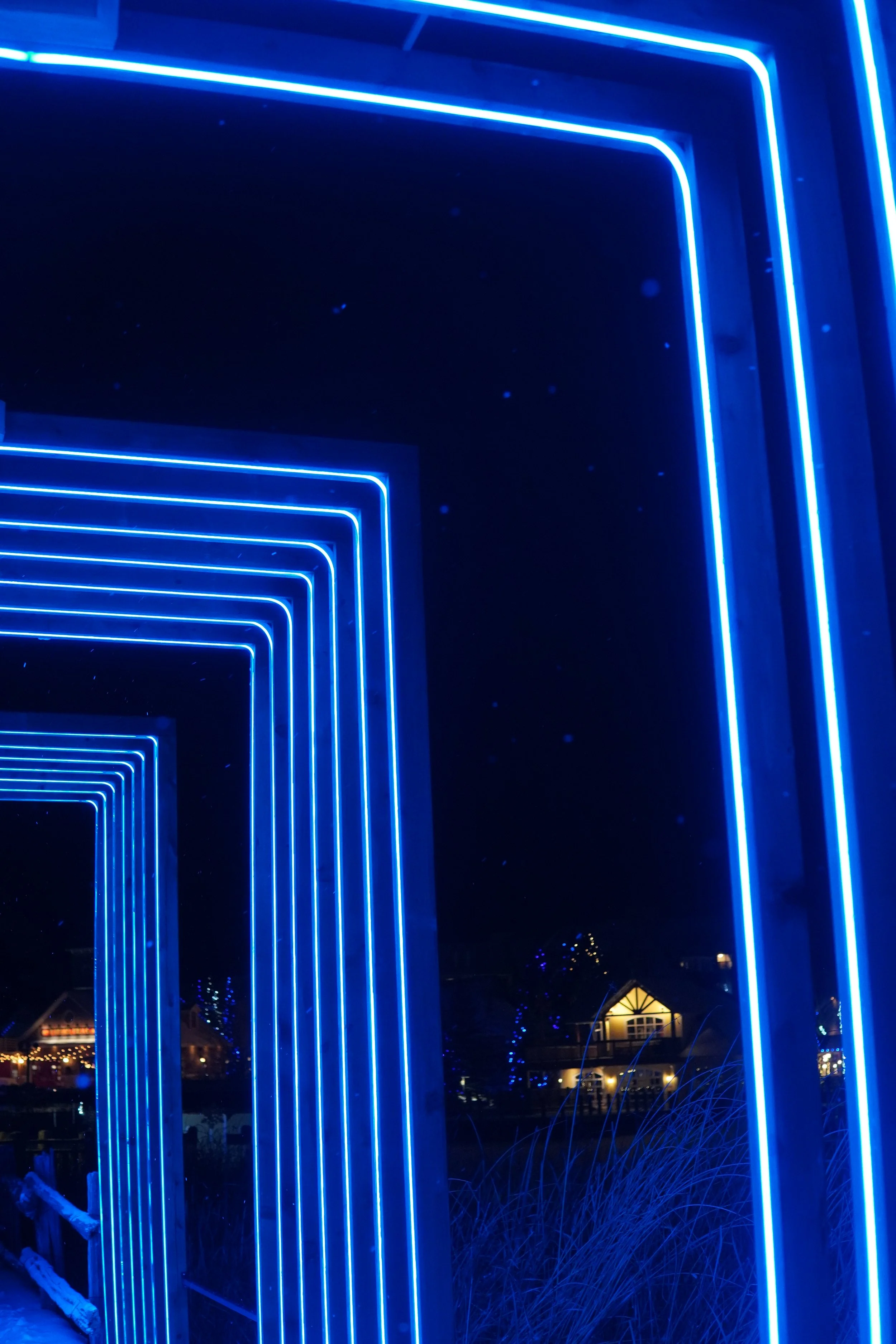 Blue neon rectangular frames creating a tunnel-like structure at night, with houses and decorative lights in the background.