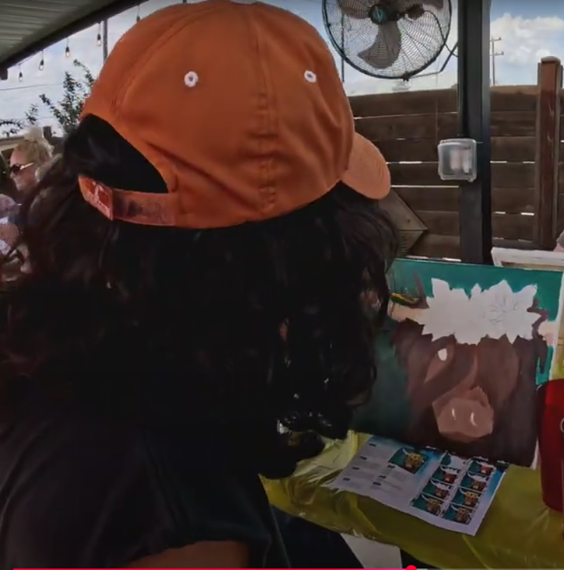 Person with long dark hair wearing an orange baseball cap, sitting at a table outdoors, viewing a painting and a pamphlet.