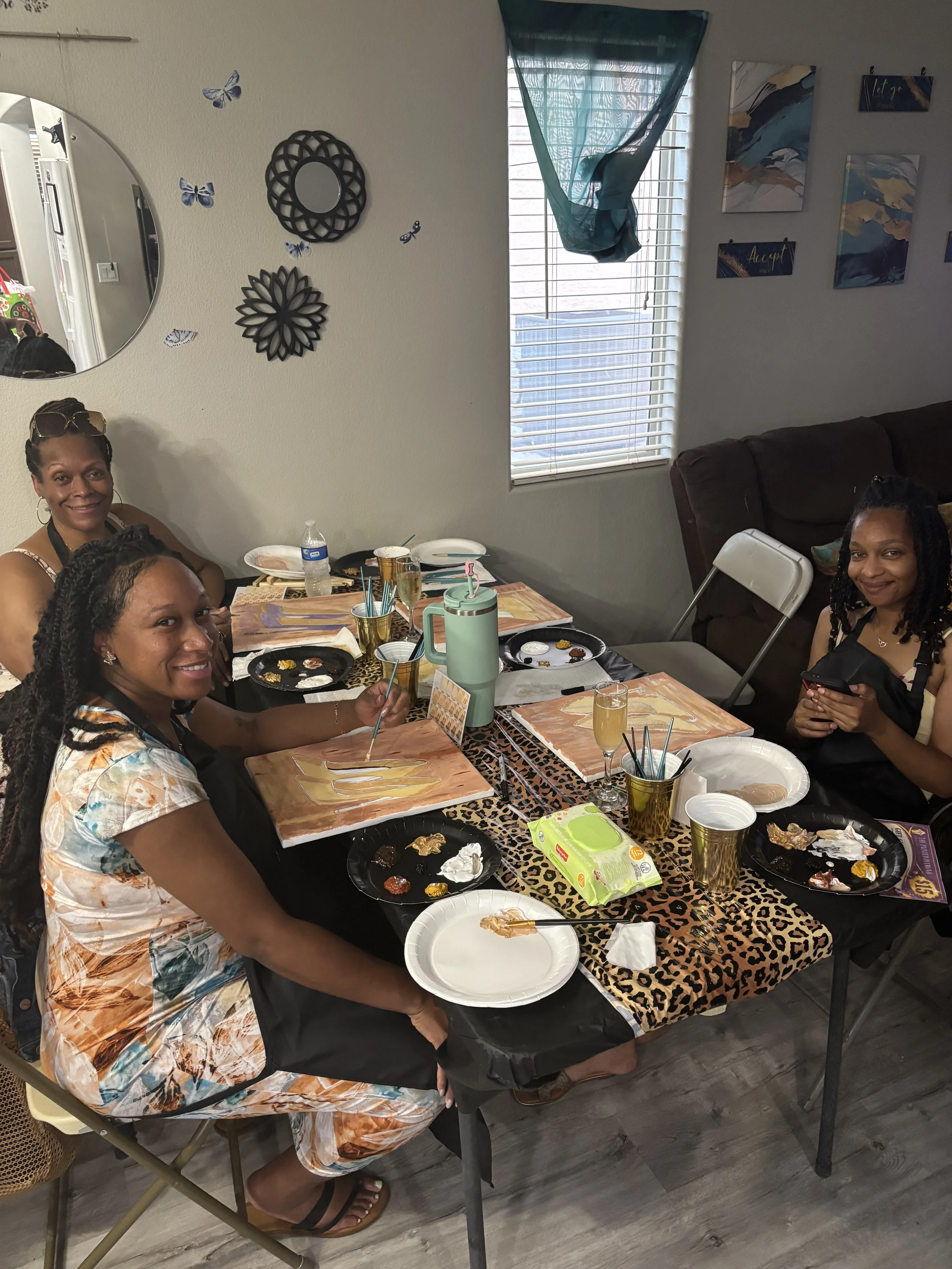 A group of women sitting around a dining table in a living room, celebrating with food, drinks, and a birthday cake with a single candle. There are plates, cups, and decorations on the table and wall art in the background.