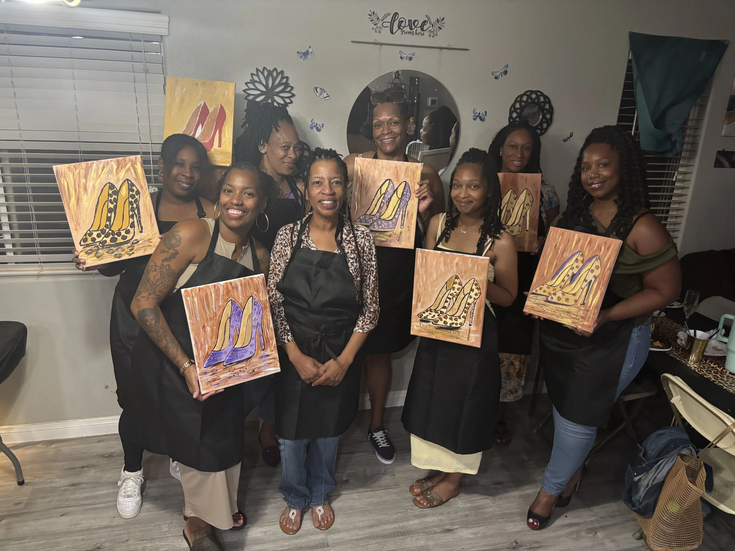 Group of women in a room holding paintings of colorful high-heeled shoes. They are smiling and wearing aprons, indicating a group painting activity.