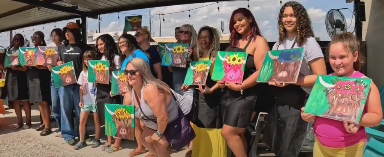 Group of women and children outdoors holding framed paintings of a cow with flowers on its head, smiling at the camera.