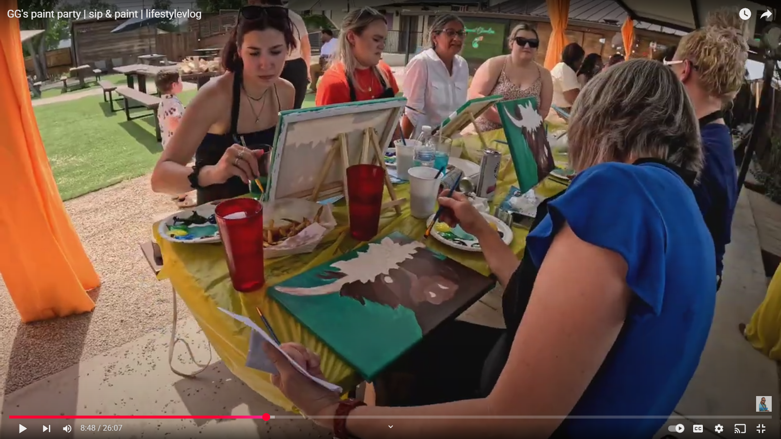 People painting animal portraits at an outdoor paint party under a canopy, with various supplies and partially finished paintings on the table.