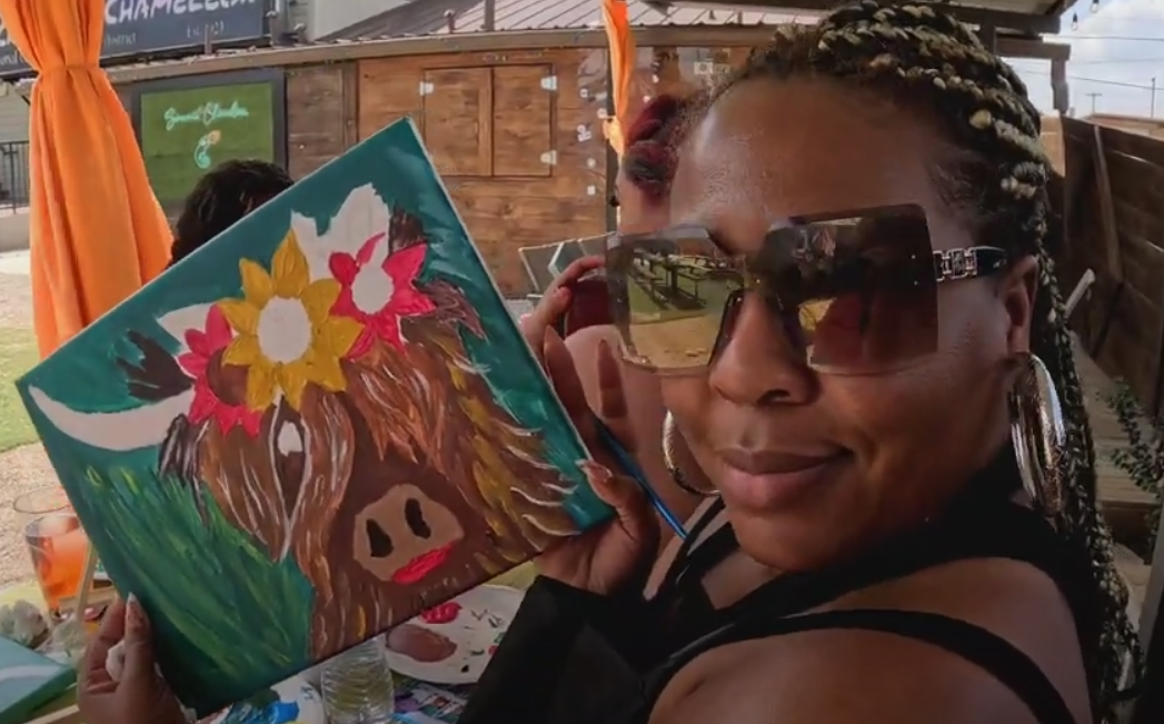 Woman with braided hair, wearing large sunglasses and hoop earrings, holding up a colorful painting of a pig with a flower crown.