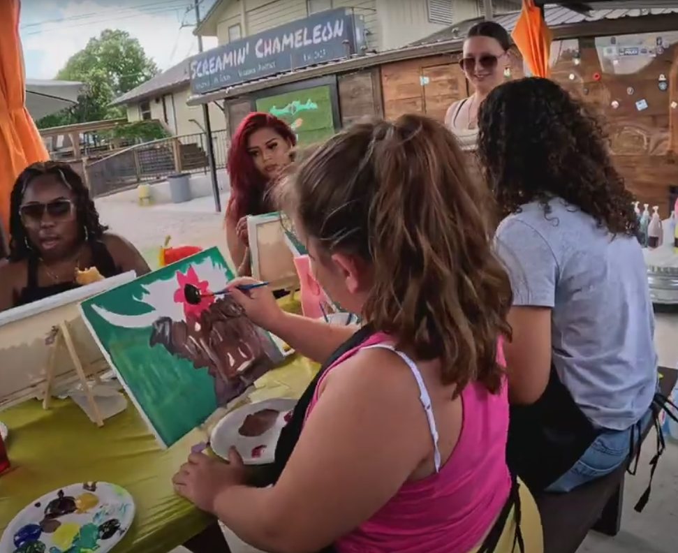A group of women and girls are gathered at an outdoor art station. One girl is painting a picture of a moose with a pink flower on its head. Others are observing or waiting their turn, and there is a sign in the background that says 'SCREAMIN CHAMELE