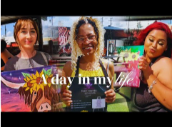 Three women standing outdoors, holding a sign that reads 'A day in my life', with stores and American flags in the background.