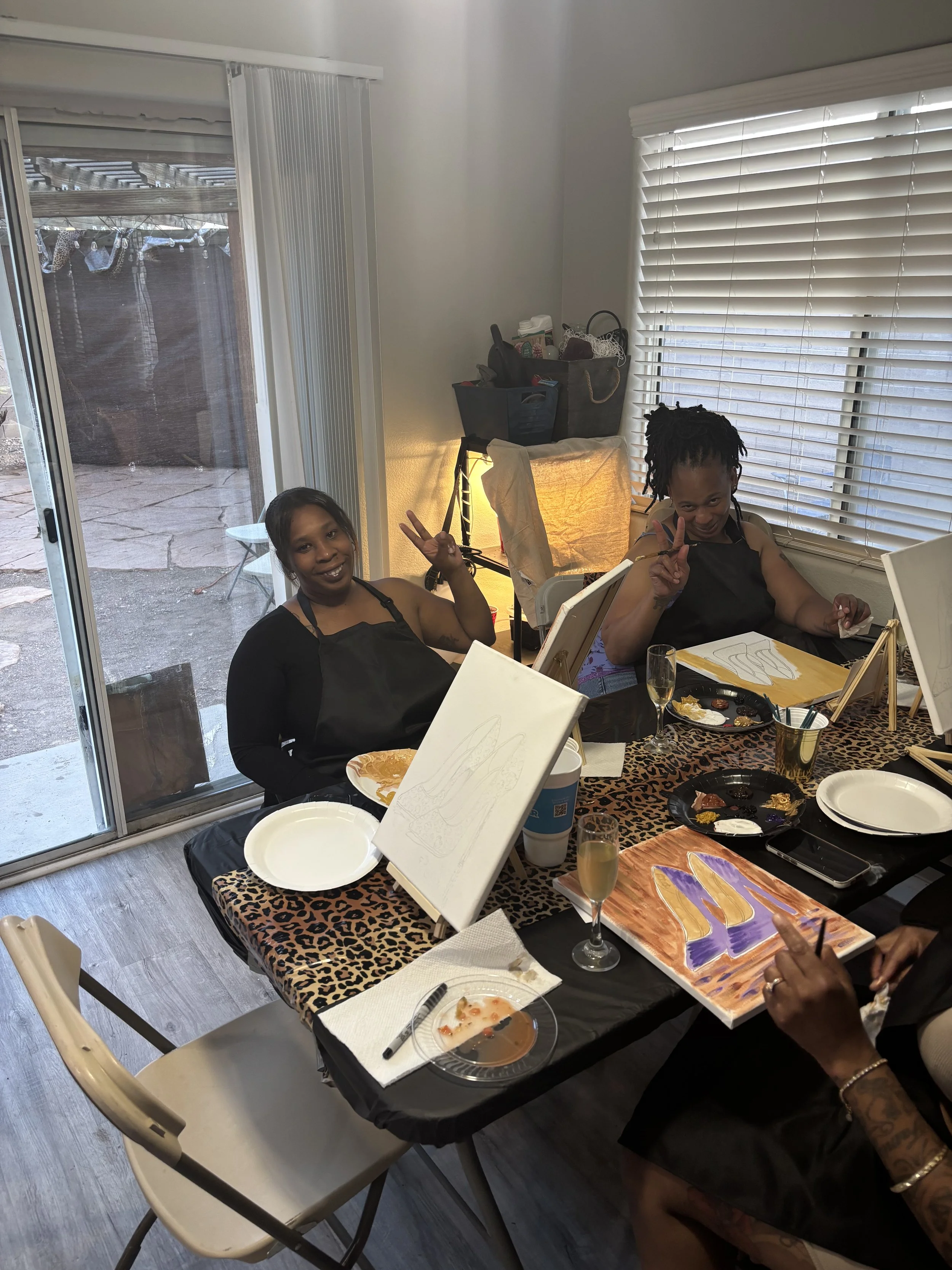 Two women sitting at a table during a painting activity, showing peace signs, with art supplies and blank canvases on the table, inside a room with large windows and blinds.