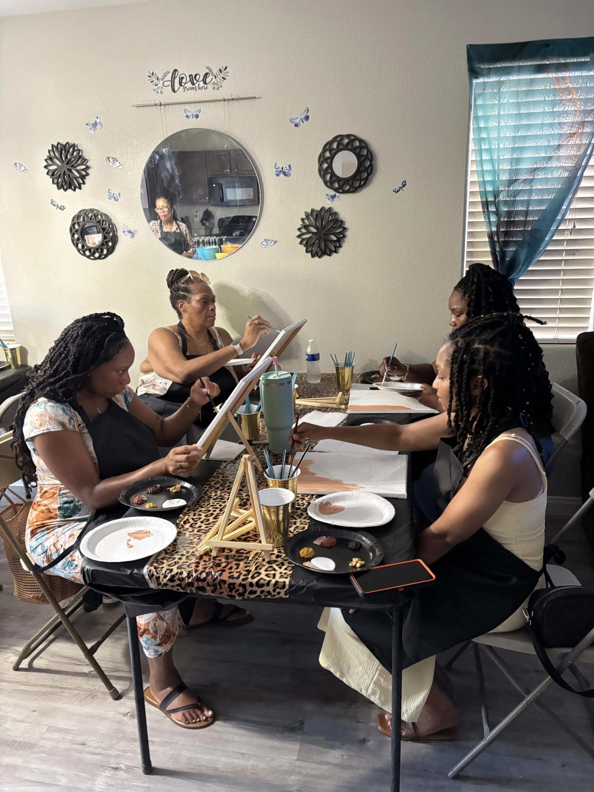 Group of women painting on canvases at a table covered with a leopard print tablecloth in a room with wall decorations and a window with blue curtains.