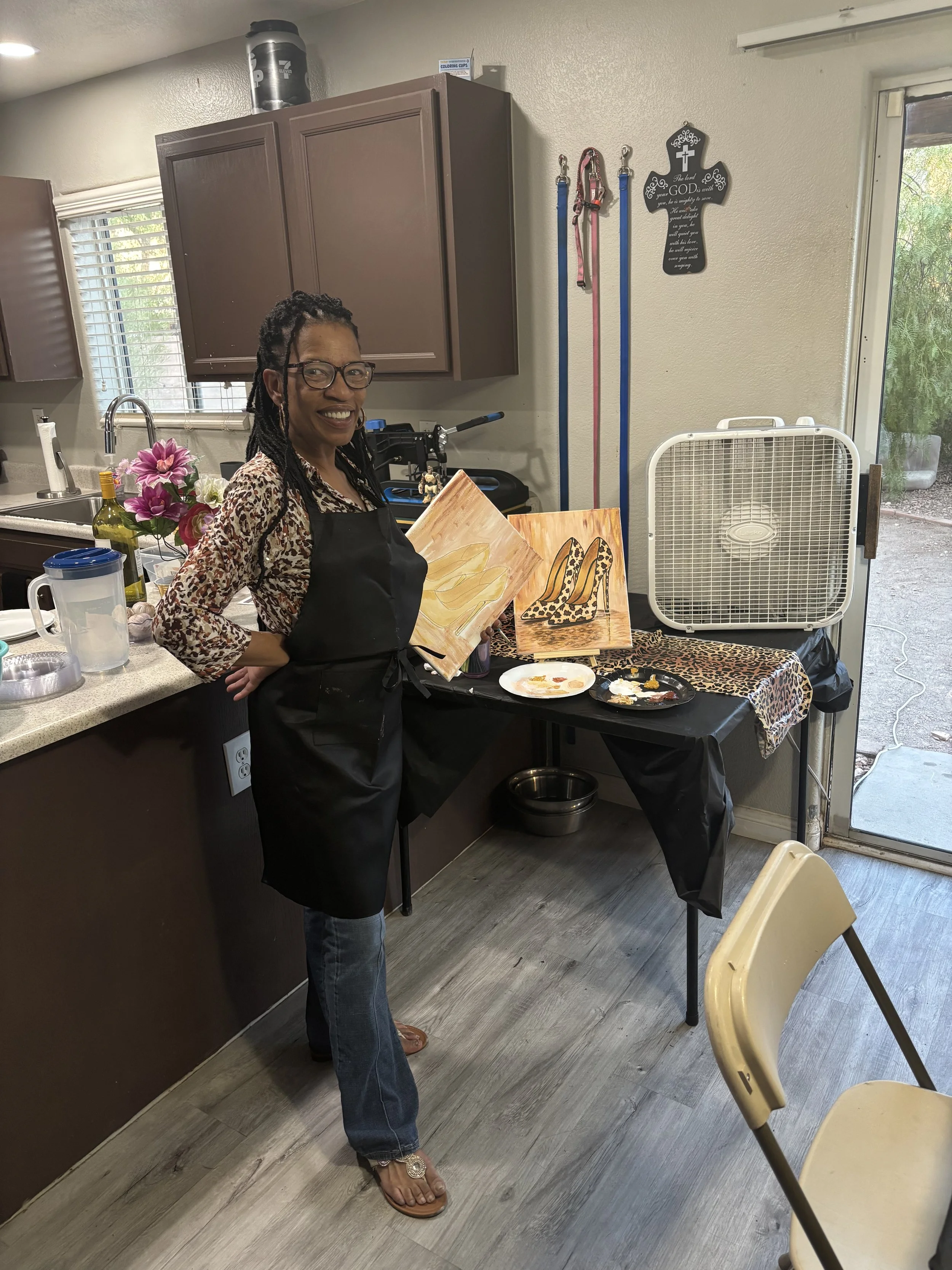A woman with glasses and braids is smiling while holding two paintings of leopard print shoes, standing in a kitchen with a table covered in art supplies and paintings. Her right hand is on her waist and she is wearing a leopard print top and a black