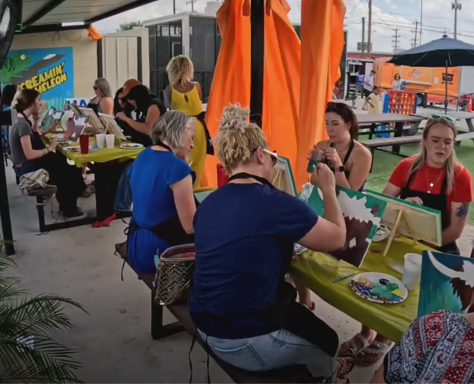 People sitting around tables at a painting event, painting on canvases, under a shelter with an orange curtain, with food and drinks on the tables, and food trucks and tents visible in the background.