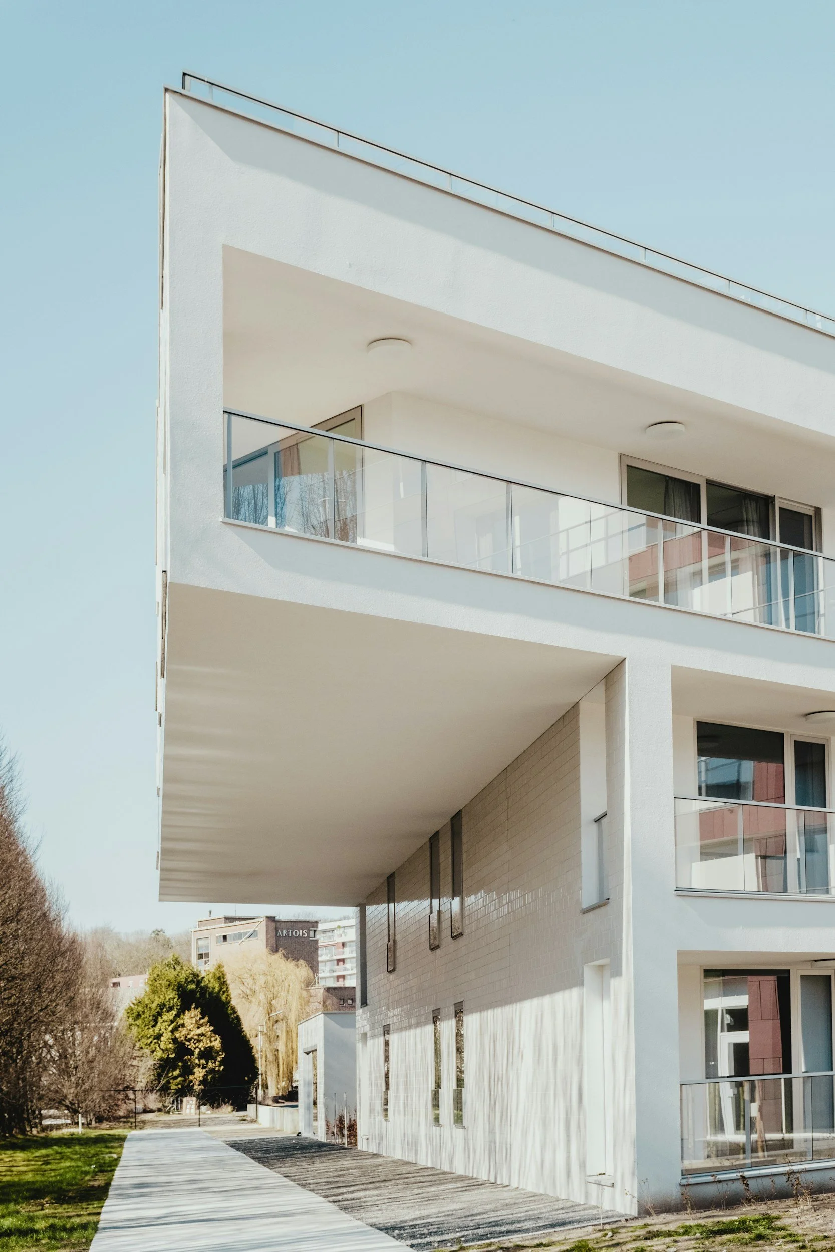 Modern white multi-story residential building with balconies and glass railings, situated at the edge of a park with trees and a walking path.
