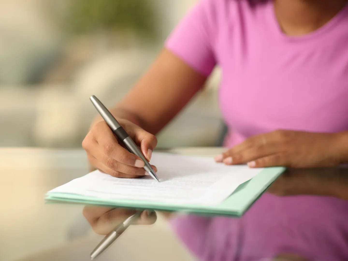 A person wearing a pink shirt is writing in a notebook with a silver pen.