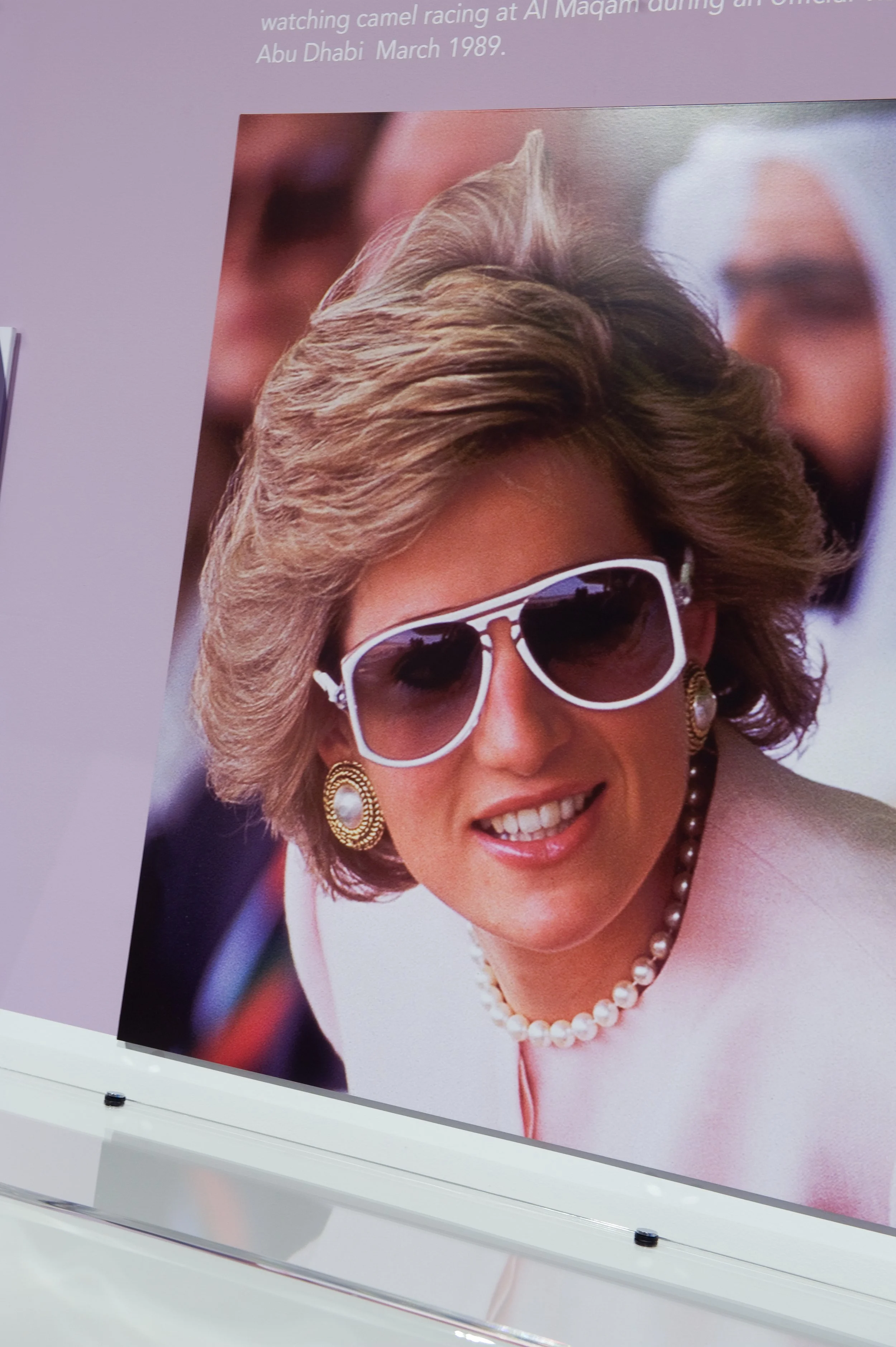 Photograph of a woman with styled hair, wearing large white sunglasses, pearl earrings, a pearl necklace, and a light-colored blazer. She is smiling and appears to be at a formal event.