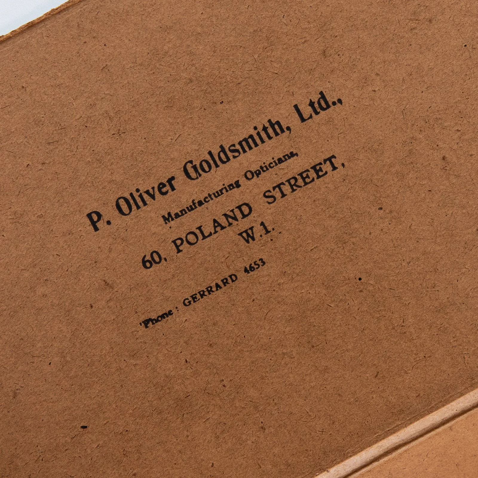 Close-up of a cardboard surface with printed text showing the name and address of P. Oliver Goldsmith, Ltd., located at 60 Poland Street, W.1, with a phone number beginning with Gerrard 4653.