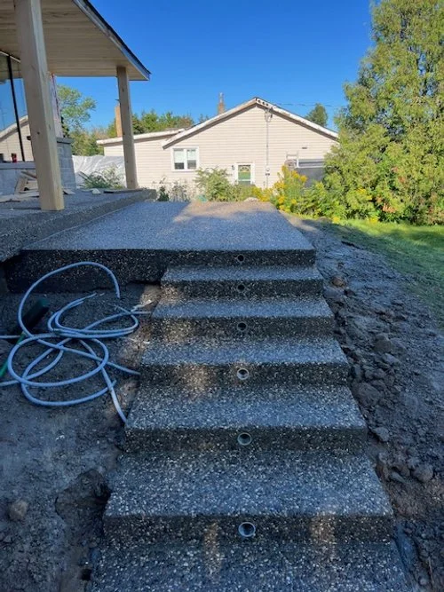 Concrete steps leading up to a covered porch in a backyard setting, with garden hose coiled on the side, surrounded by grass and trees.