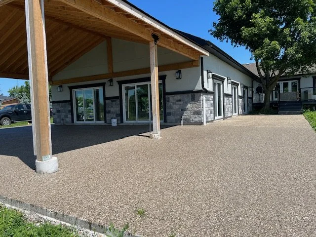 Outdoor patio area with concrete flooring, wooden beams, and glass sliding doors, adjacent to a building with stone accents and surrounded by greenery.