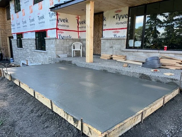 Newly poured concrete patio in front of a partially constructed house with stone and Tyvek outer walls.