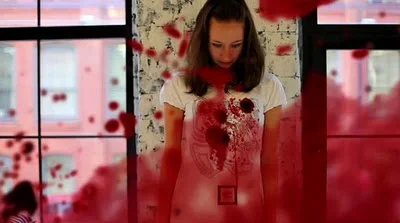 A woman standing inside a room with large windows, bloodstains splattered on the windowpanes and her shirt, looking down with a serious expression.