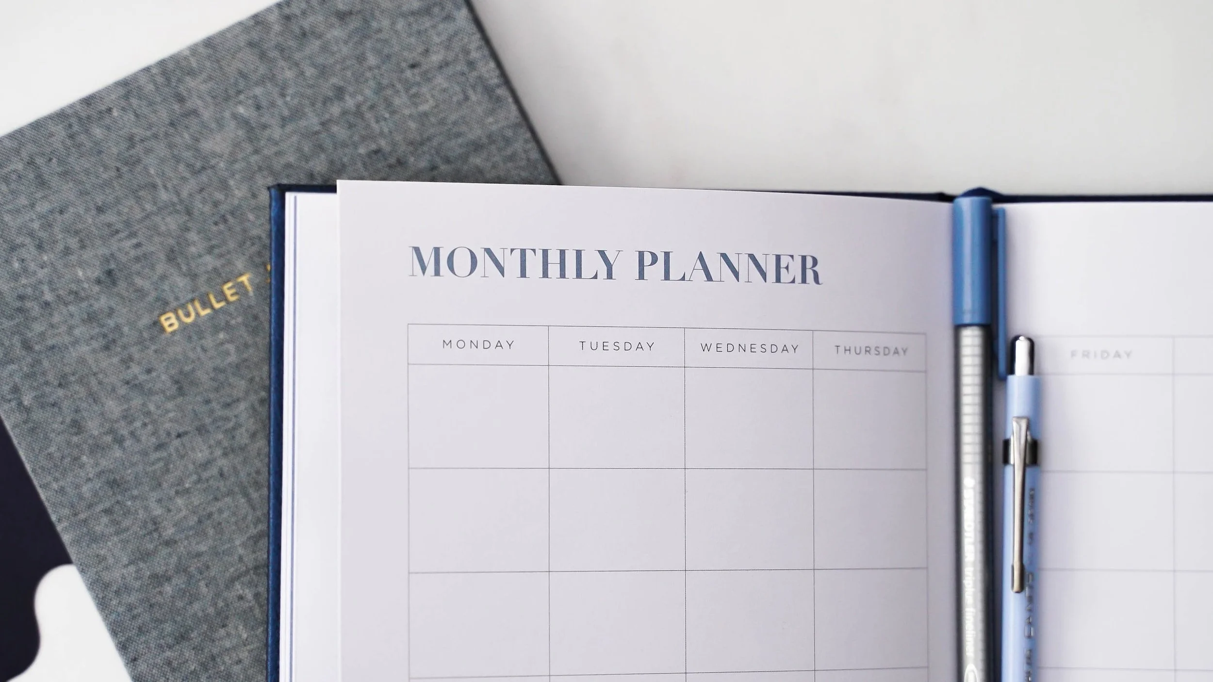 Open monthly planner with a blue pen clipped to on the right side, resting on a gray Bullet journal notebook.