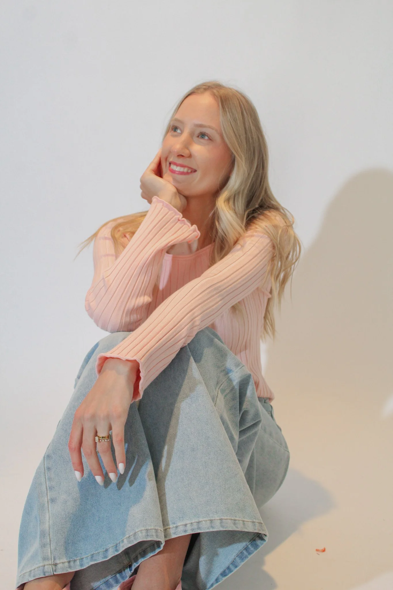 A young woman sitting on the floor, smiling, with one hand resting on her cheek. She is wearing a pink, long-sleeved, pleated top and light blue jeans.