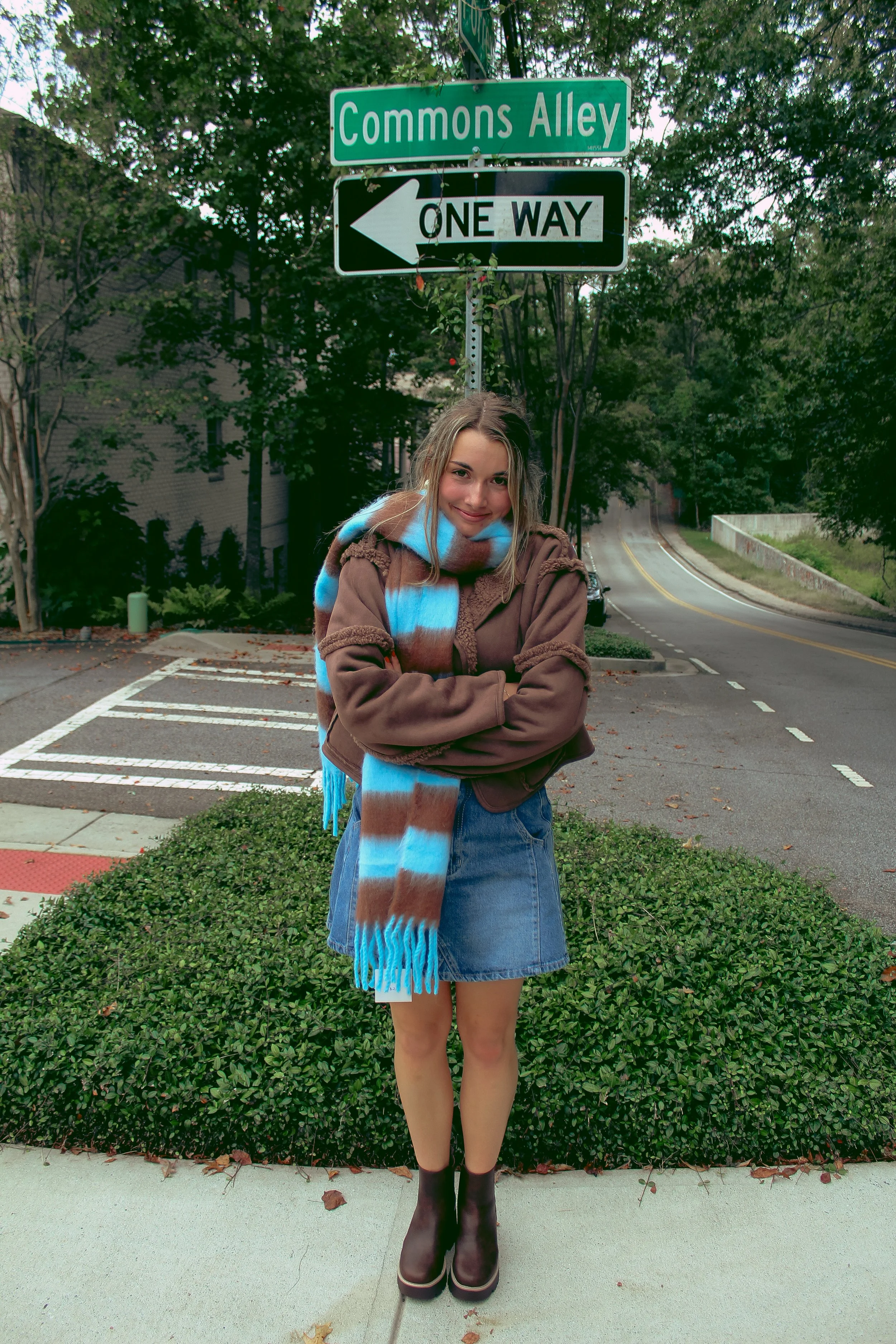 A young woman with a scarf, jacket, denim skirt, and boots standing on a sidewalk in front of street signs indicating 'Commons Alley' and 'One Way' in a residential area with trees.