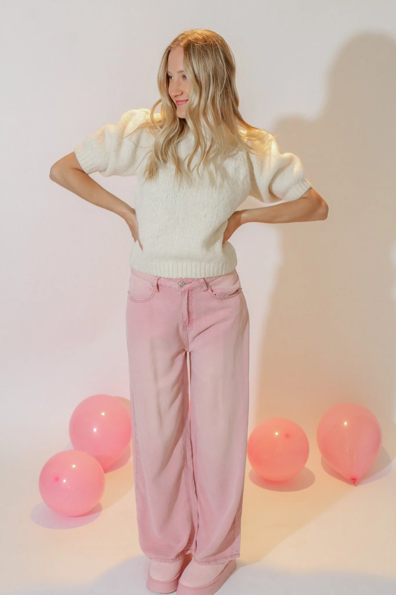 A woman in a cream sweater and pink wide-leg pants standing with her hands on her hips, surrounded by four pink balloons against a plain background.