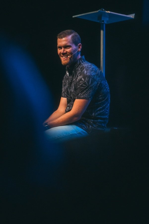 A man with short hair and a beard, smiling and wearing a patterned short-sleeve shirt, sitting on stage with a black background and a blue spotlight shining on him.