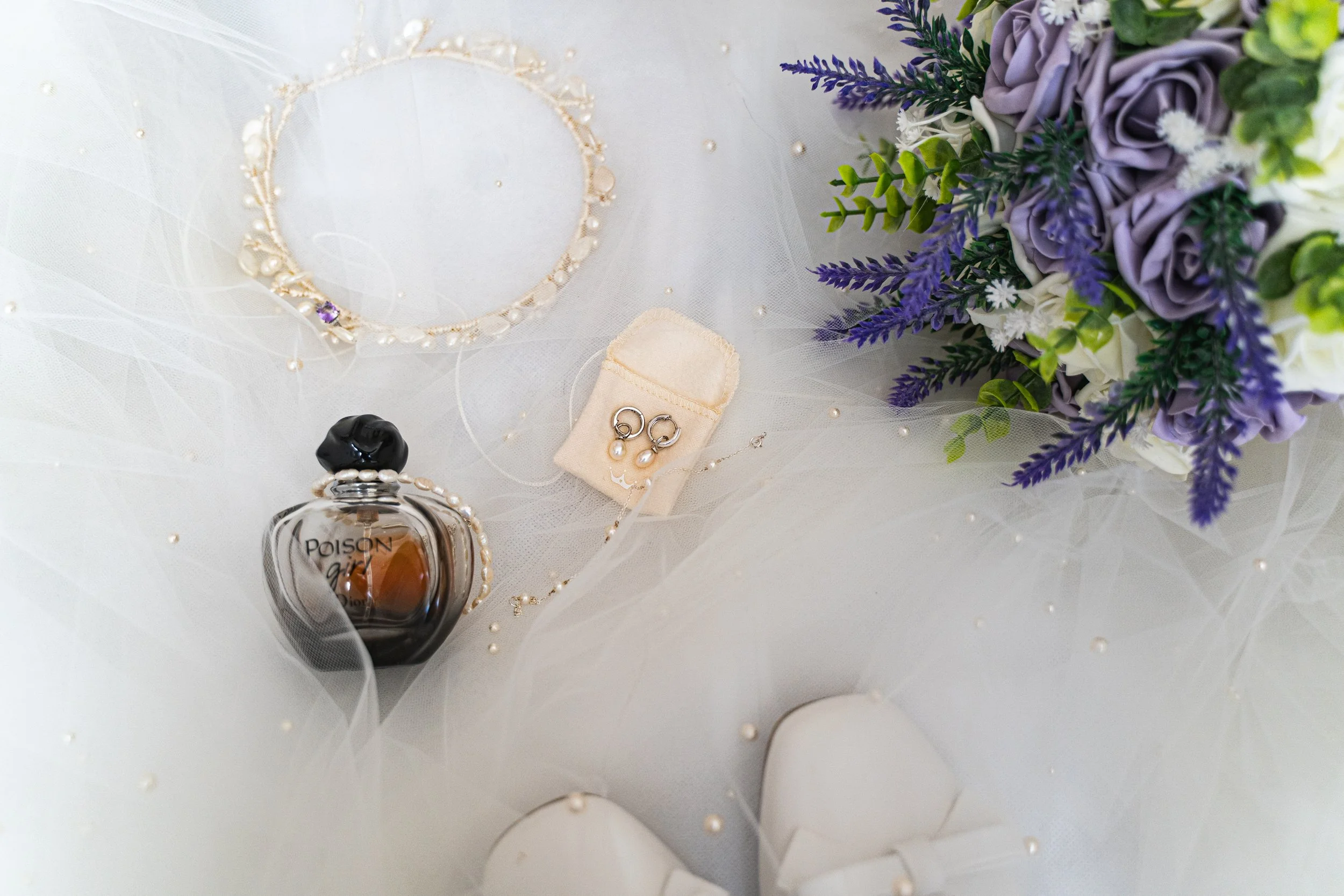 A flat lay of floral jewelry, a perfume bottle, and jewelry box on sheer fabric with faux pearl accents, next to a purple and green flower bouquet.