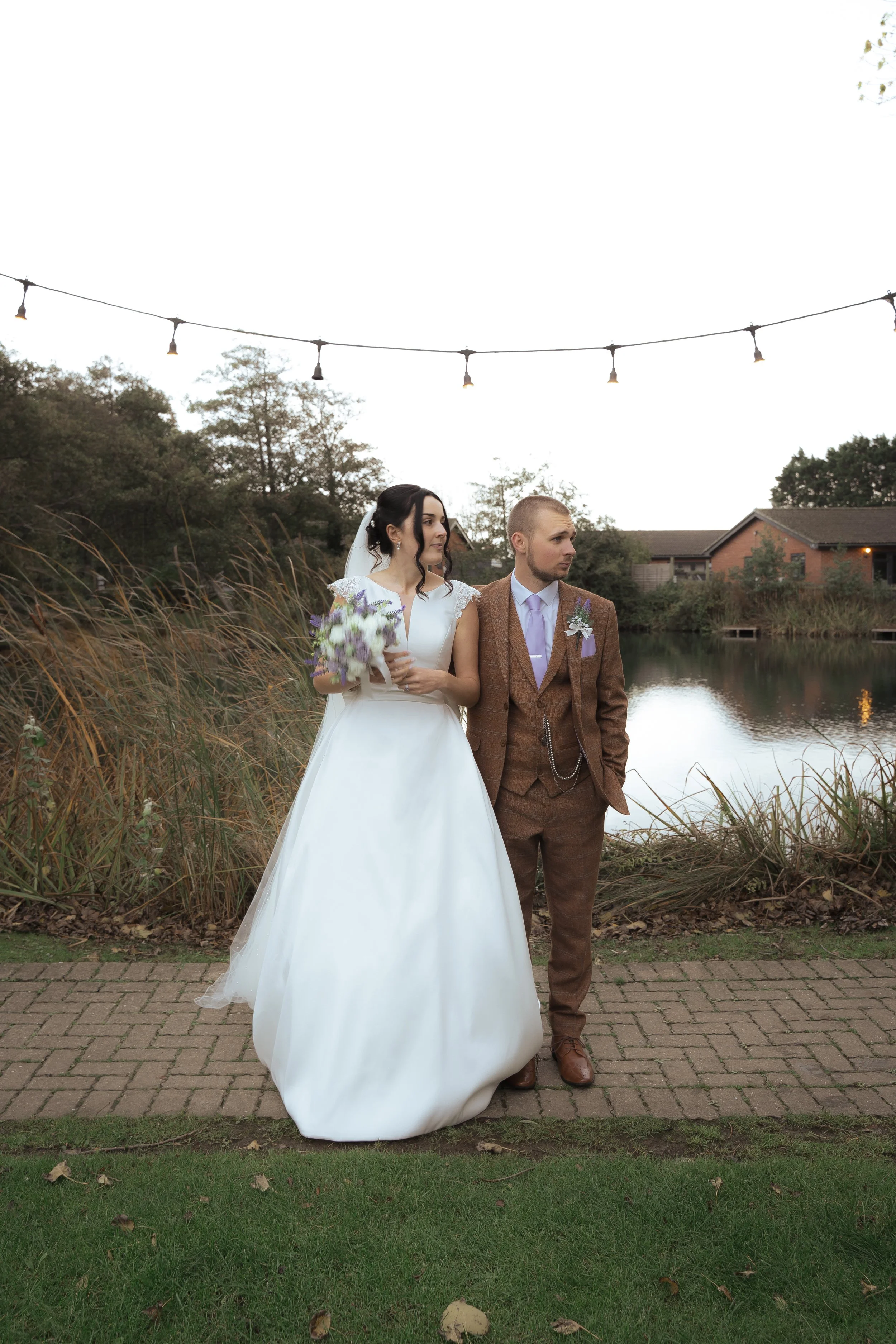A bride and groom stand outdoors near a pond, with tall grass and trees behind them, under string lights on an overcast day.