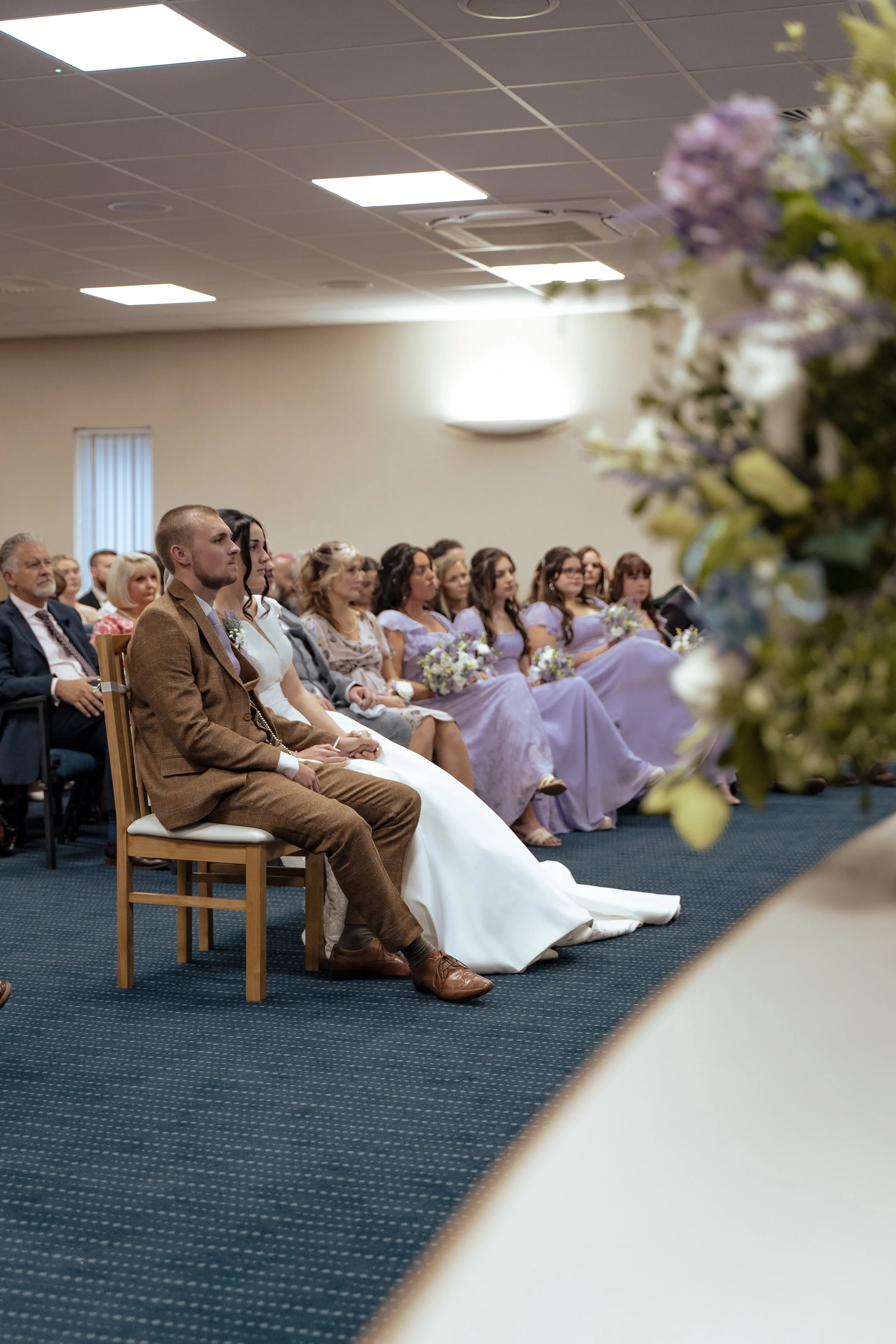 Wedding ceremony with guests sitting in rows, including a bride in a white gown and a groom in a brown suit, seated on wooden chairs, with purple flowers visible in the foreground, inside a room with a suspended ceiling, fluorescent lights, and beige