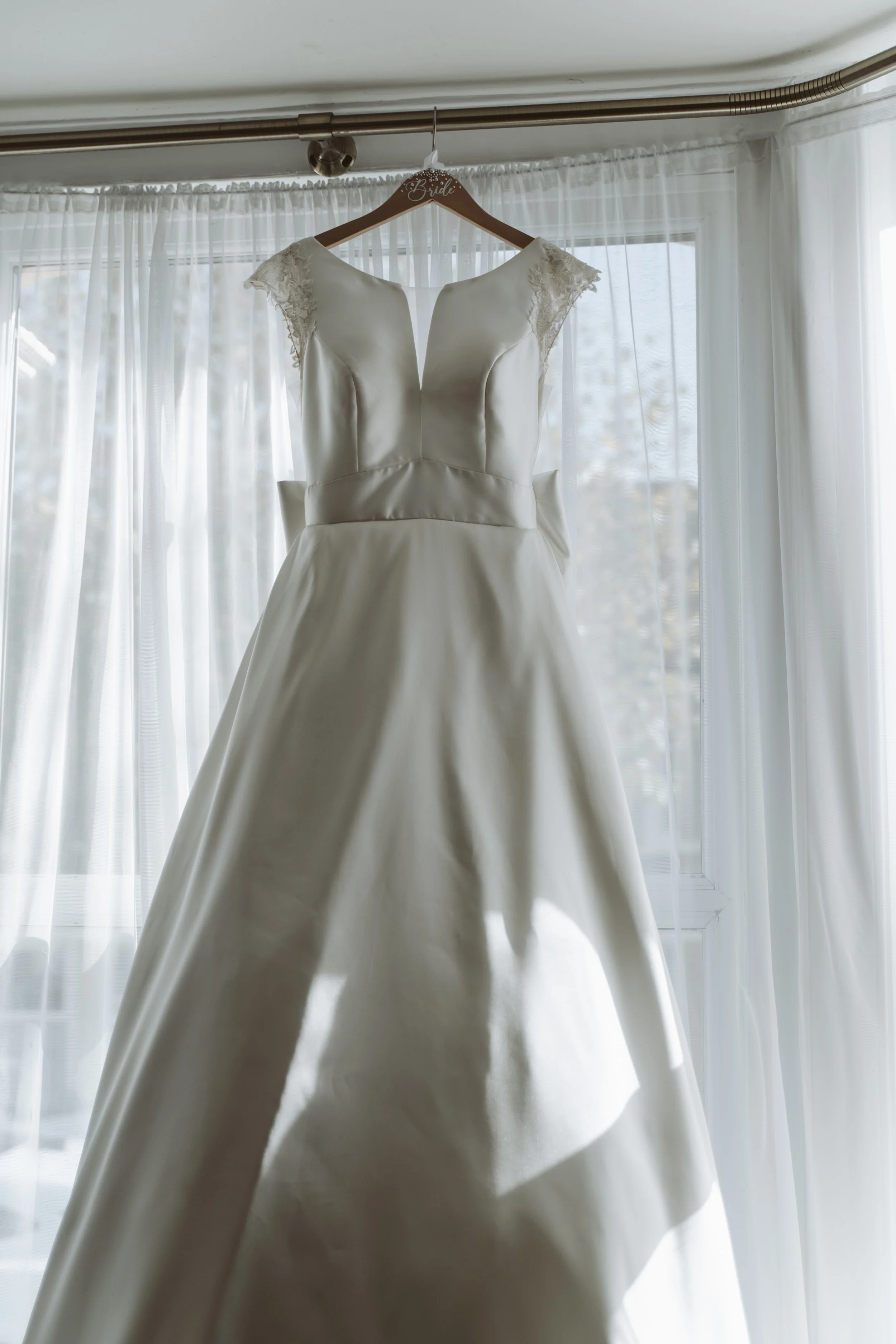 White wedding dress hanging on a wooden hanger labeled 'Bride' in front of a window with sheer white curtains.